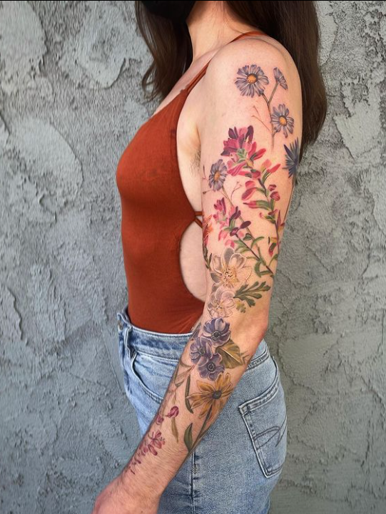 A woman with floral tattoos on her upper arm and forearm, wearing a brown sleeveless top and high-waisted jeans, standing against a textured gray wall.