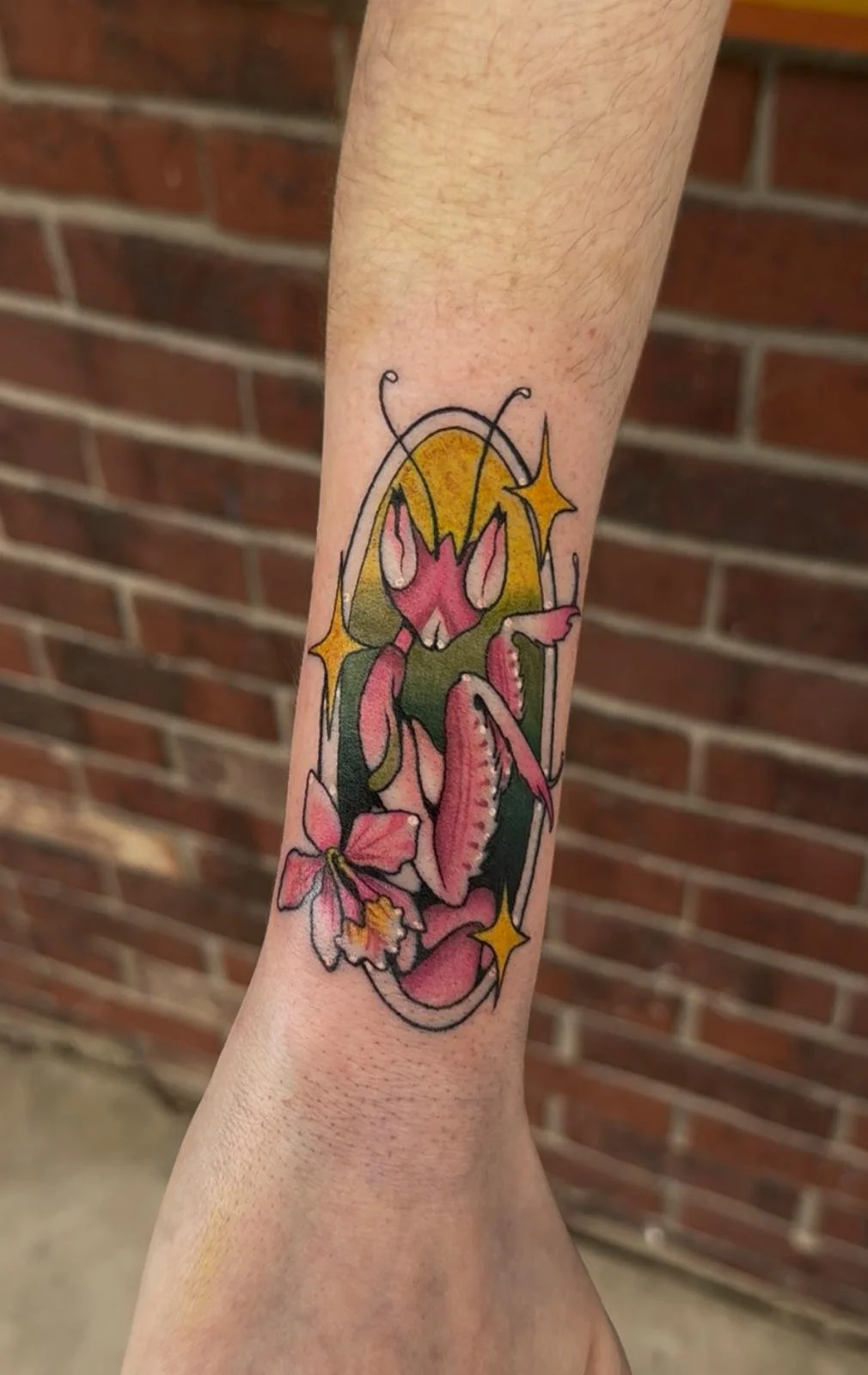Colorful tattoo of a praying mantis with pink and green details, surrounded by yellow sparkling stars and pink flowers, on a person's forearm.