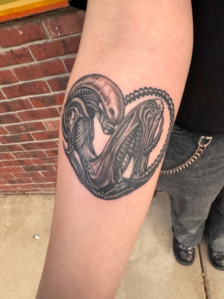 Black and gray tattoo of a science fiction-style alien creature on a person's upper arm.