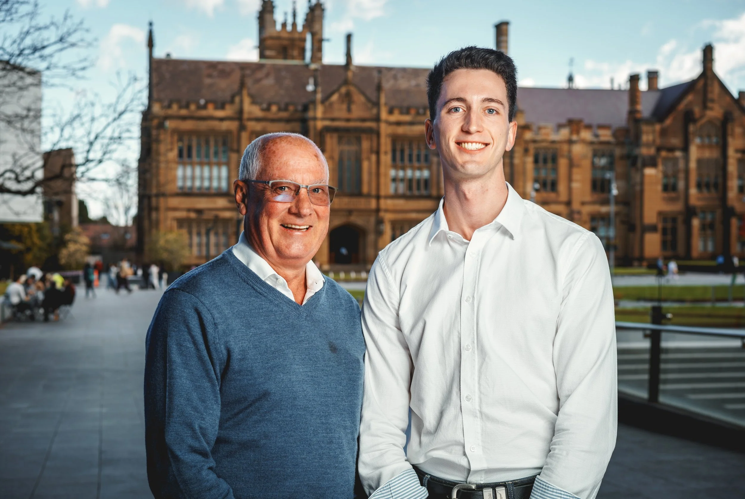 Jan-Willem and Jett van der Wallen standing in front of the University of Sydney. The older man on the left wears glasses and a blue sweater, the younger man on the right wears a white shirt.