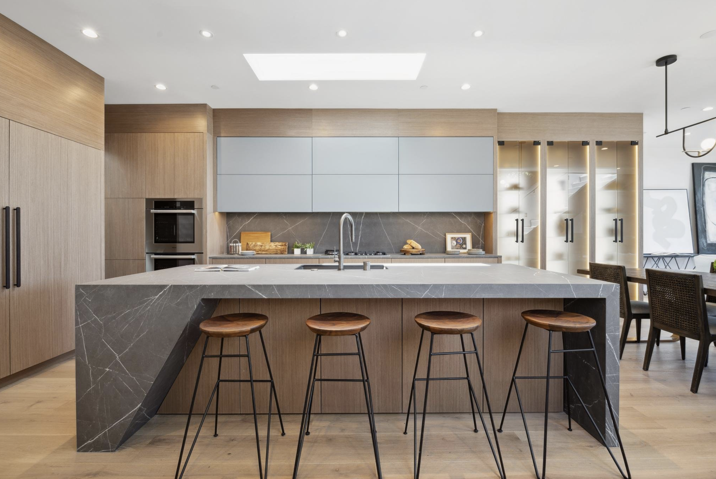 Modern custom kitchen with light wood cabinetry, gray upper cabinets, glass-front storage, and a stone waterfall island