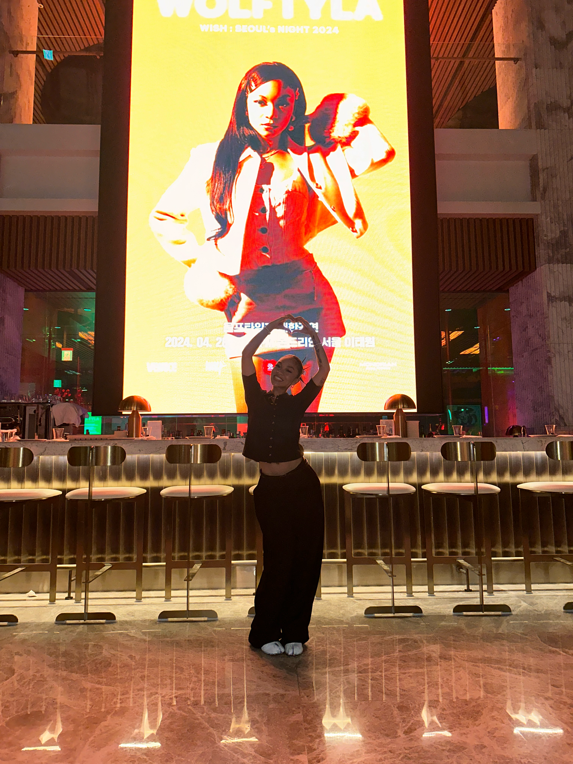 Person standing in front of a large promotional poster in a modern bar, featuring a performer on an orange background.