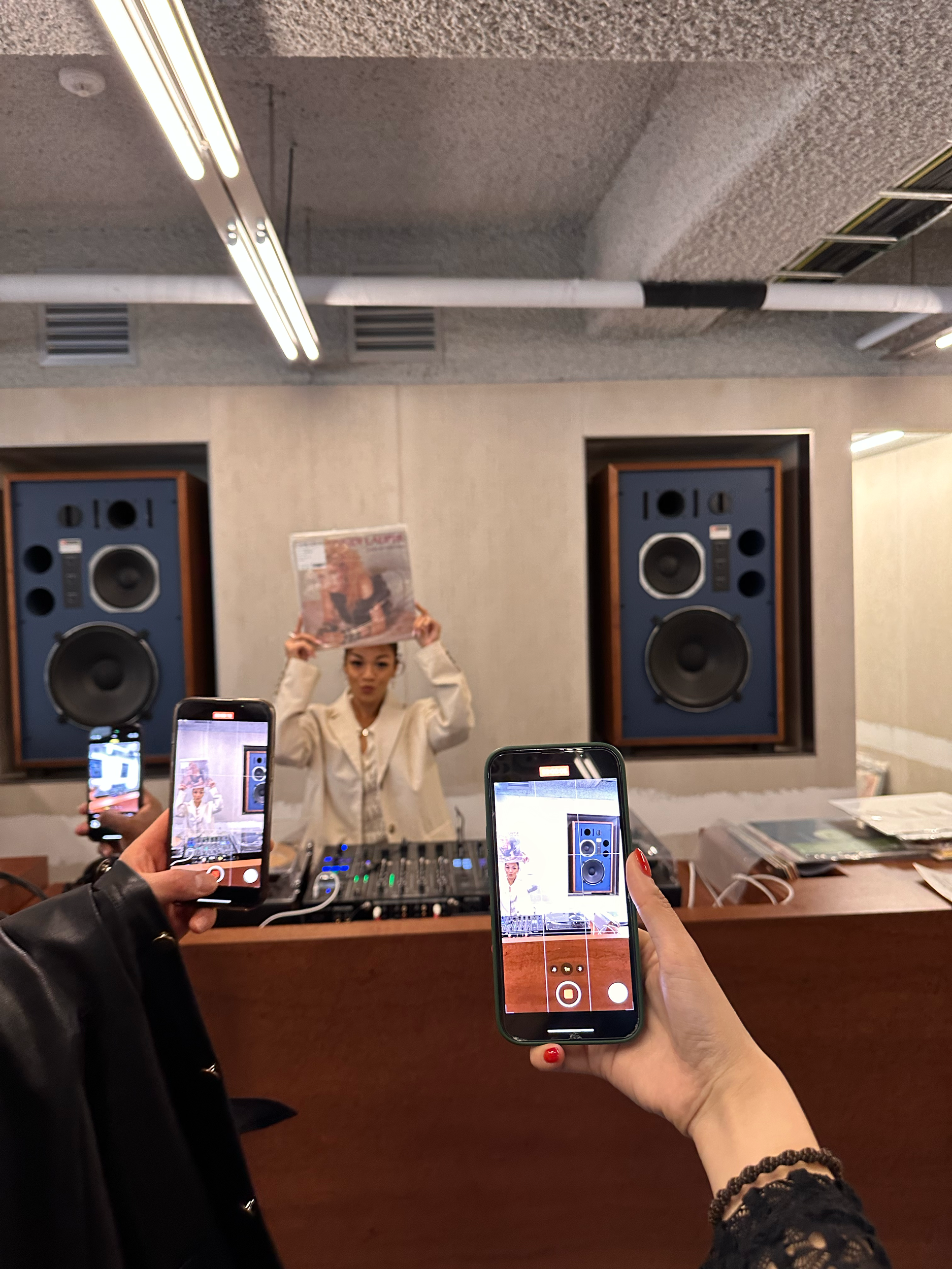 A person in a studio holding up a magazine while being photographed by others. Visible are a DJ setup, loudspeakers, and people using smartphones to take pictures. The studio has a modern design with panel walls and ceiling lights.