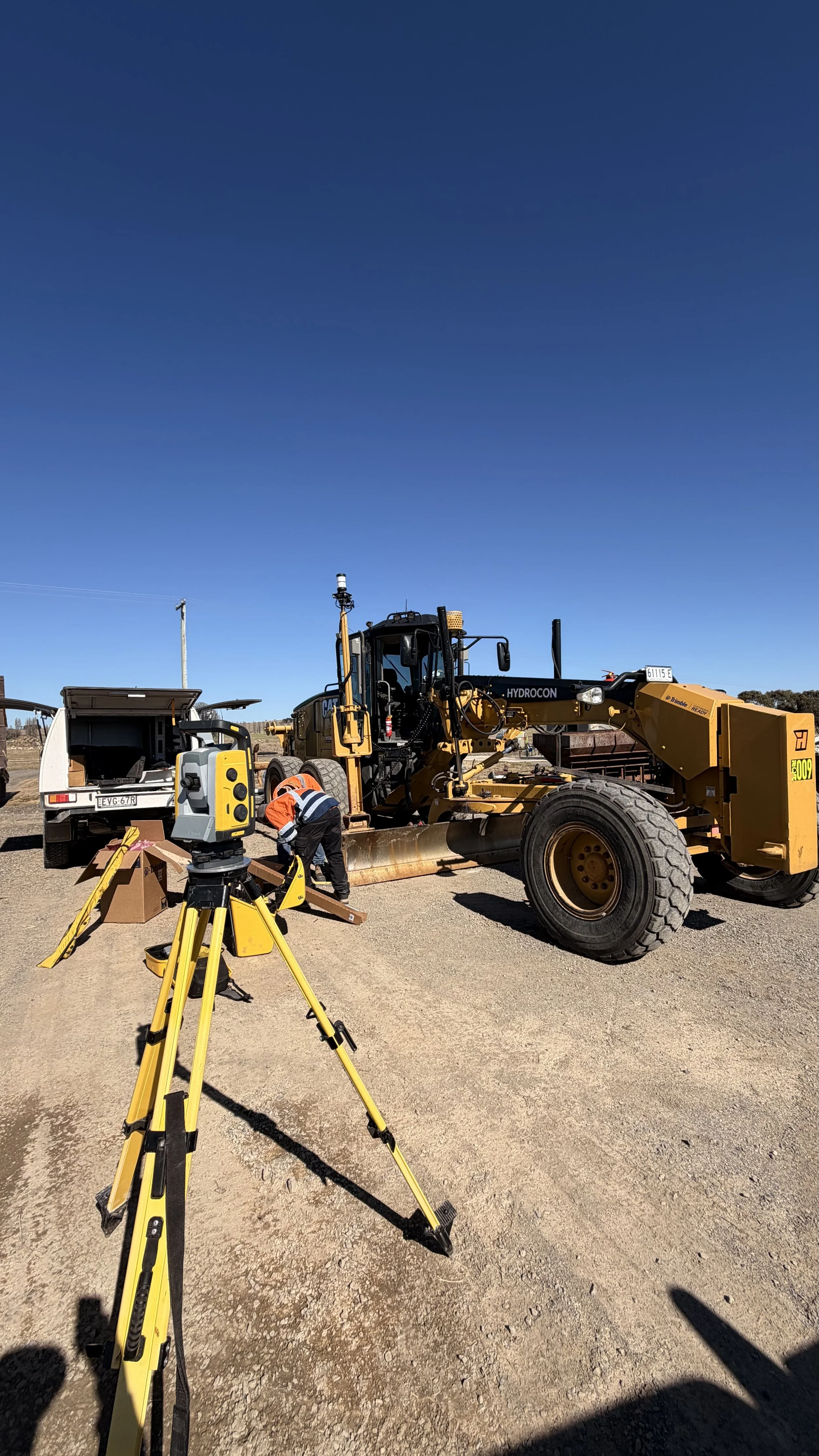 Construction workers operate surveying equipment with a large bulldozer on a dirt site under a clear blue sky.