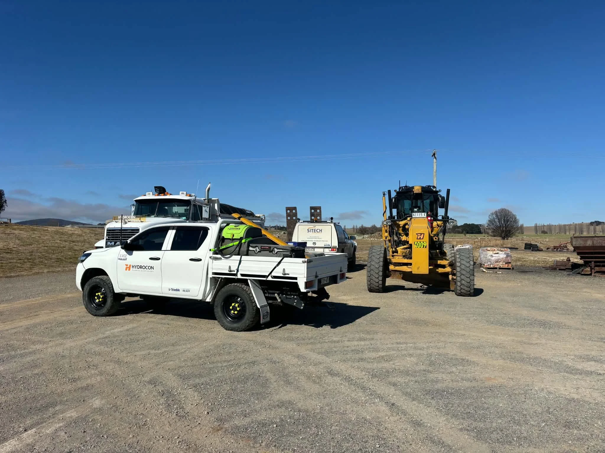 Construction site with a white Hydrocon truck, a yellow heavy-duty machine, and construction materials on dirt ground under a clear blue sky.