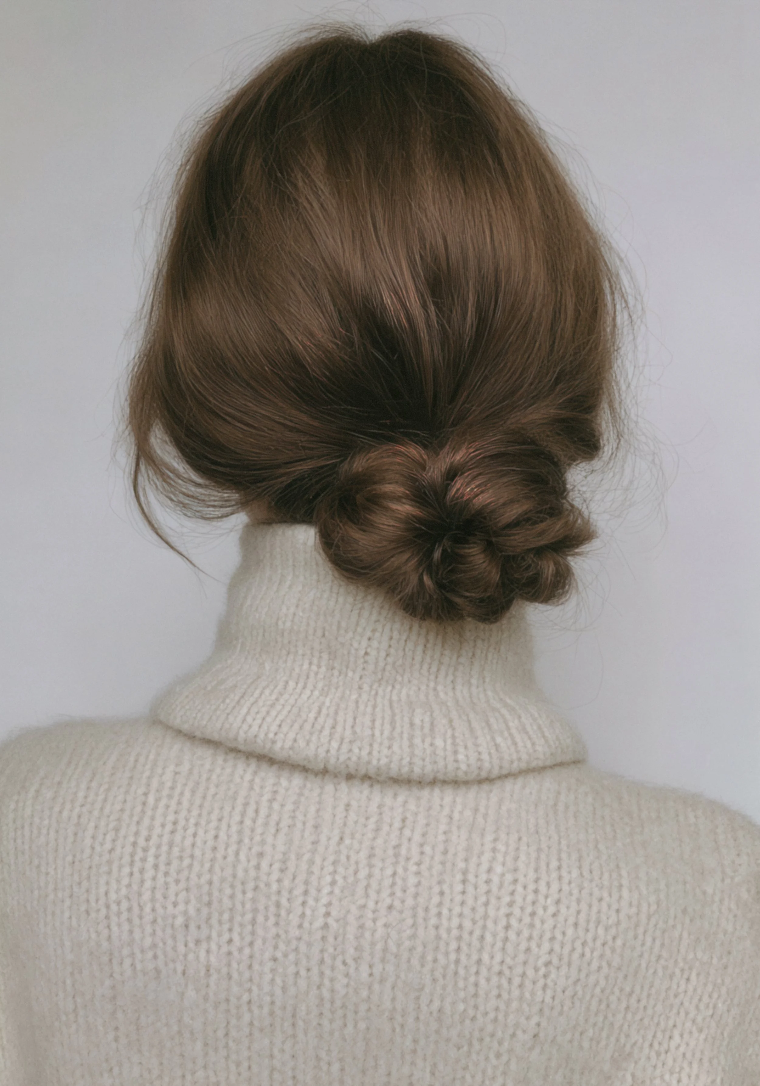 A woman with brown hair styled in a low, twisted bun, wearing a cream-colored turtleneck sweater, against a plain, light background.