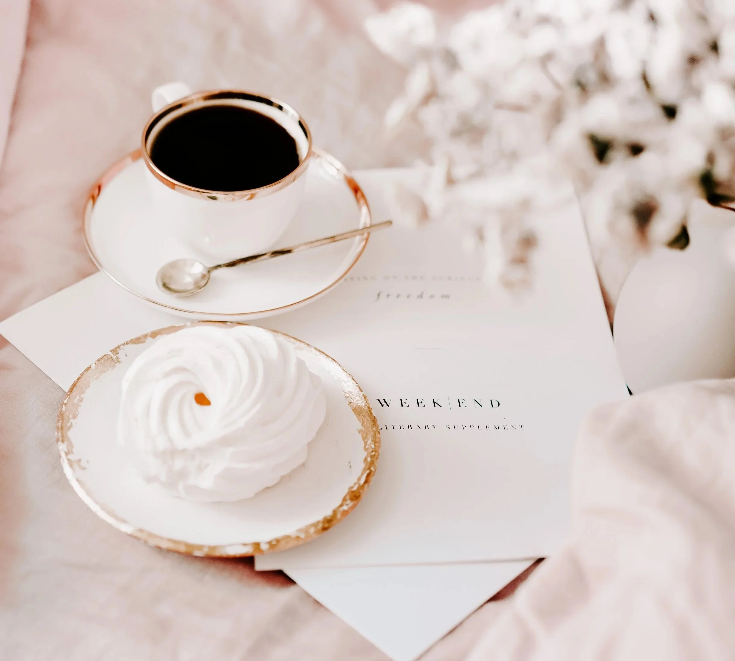 A cup of black coffee on a saucer with a silver spoon, a meringue dessert on a gold-rimmed plate, and some white flowers in the background, placed on a soft pink surface with papers and a pen.