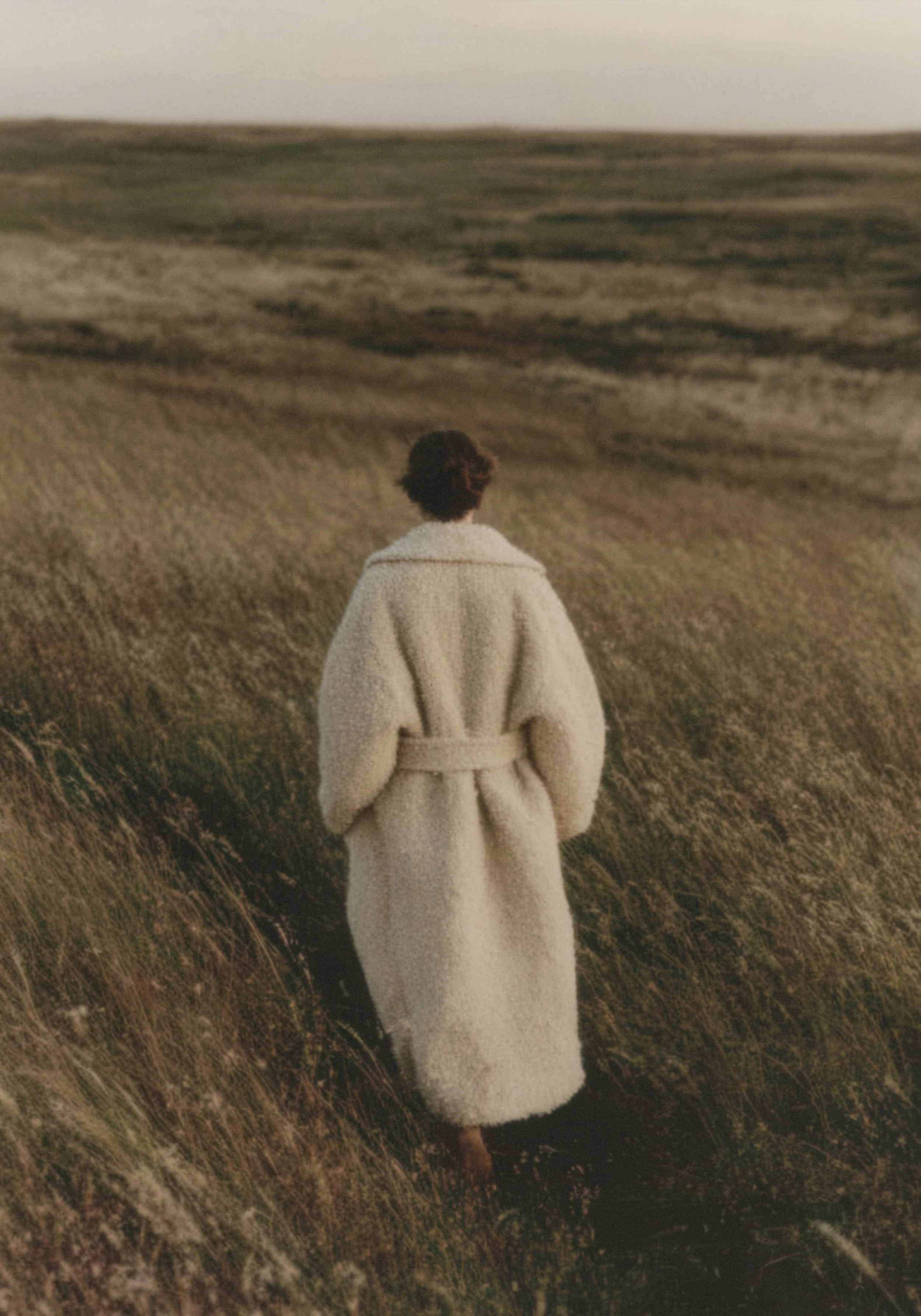 Person with curly hair wearing a white coat walking through a field of tall grass or wheat, with hills in the background during dusk or dawn.