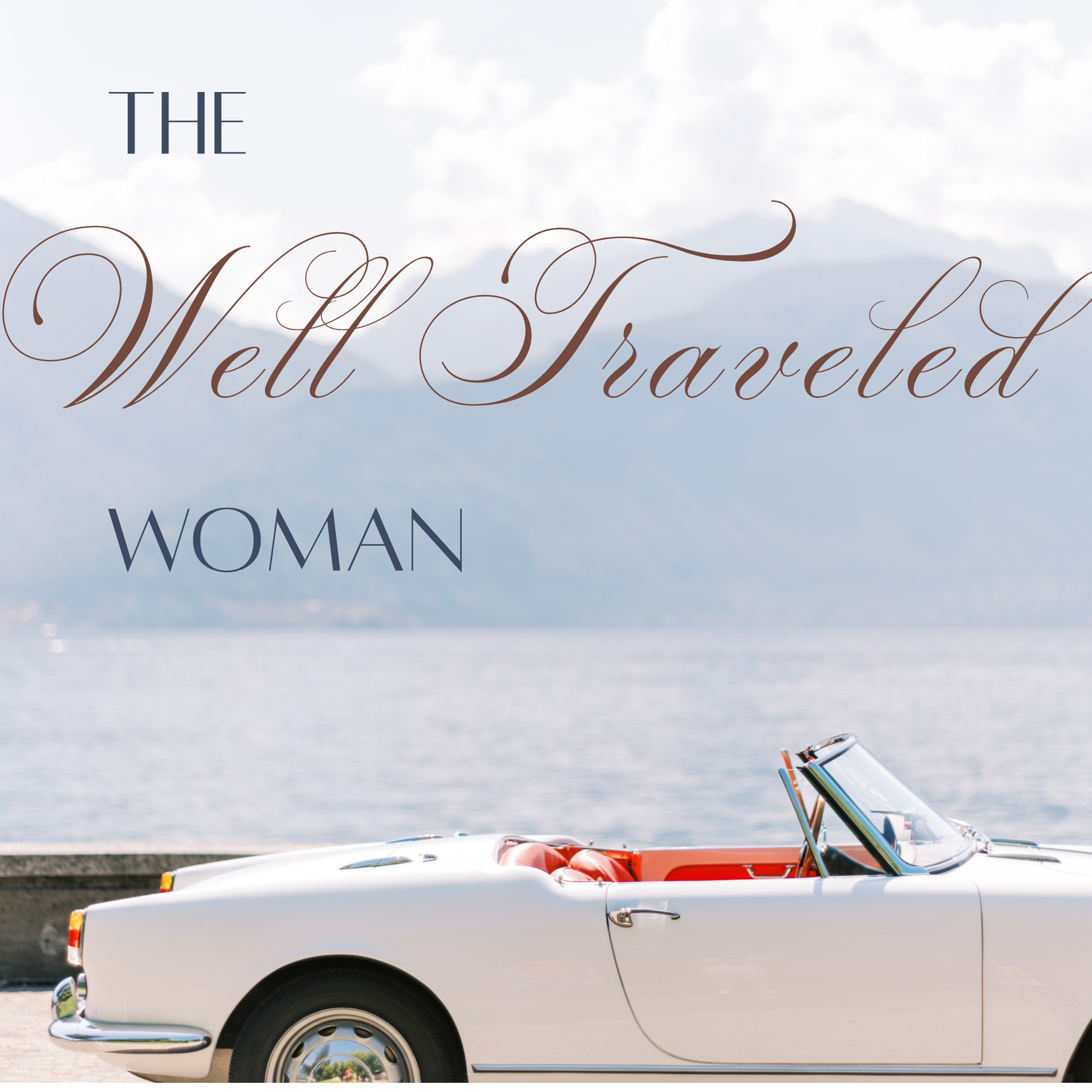 A white vintage convertible car with a red interior parked by a body of water with mountains in the background. There are some seat belts and a folding windshield visible.
