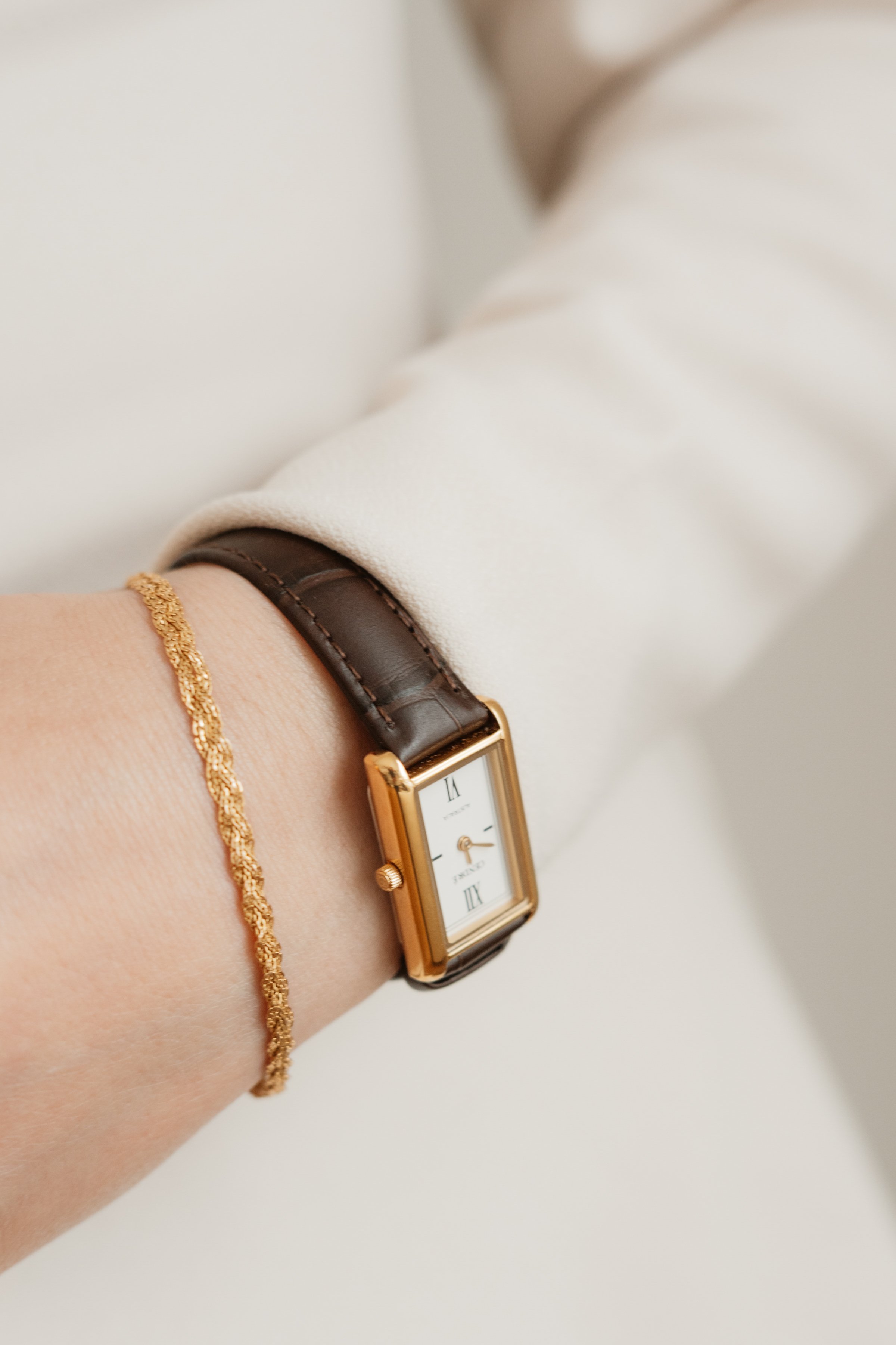 Close-up of a person's wrist showing a gold bracelet, a watch with a rectangular face and leather strap, and a brown leather strap on white clothing.