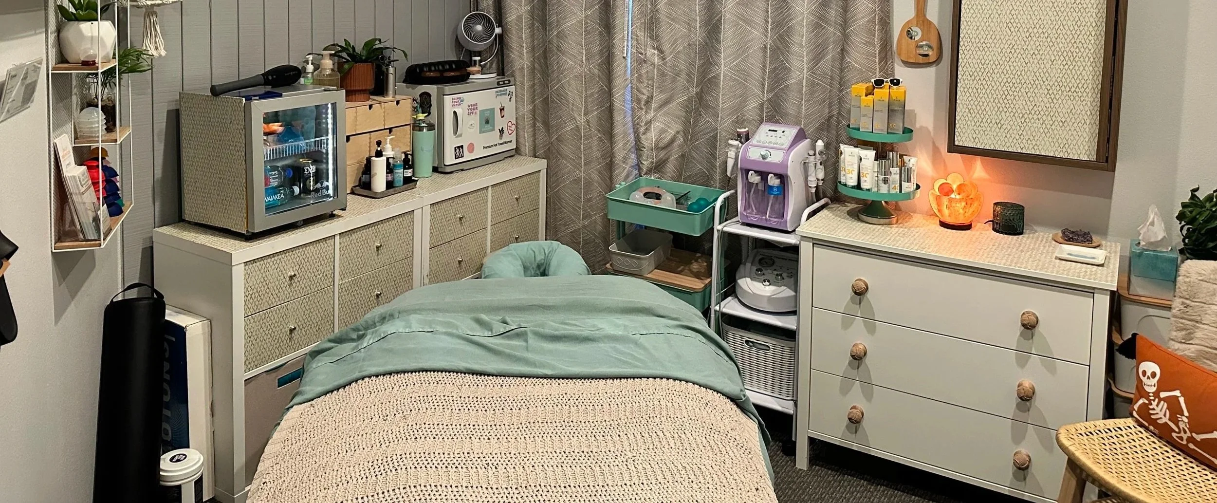 A cozy room containing a bed with a beige crocheted blanket and a green bedsheet, a white dresser with wooden knobs, a mini refrigerator, a sterilizer, and various skincare and health devices on the furniture. The room is decorated with potted plants, a glowing Himalayan salt lamp, and a skull-themed pillow. Curtains cover the window, and the space has a gentle, relaxing ambiance.