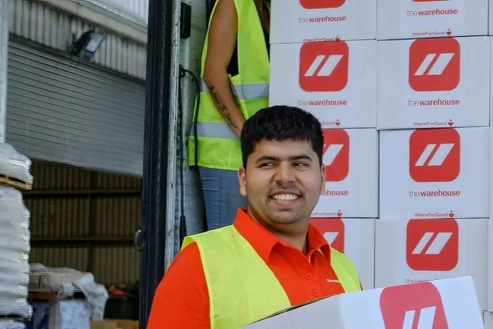 A man wearing a red shirt and yellow safety vest smiling, standing in front of stacked boxes with Warehouse logo, near an open truck.