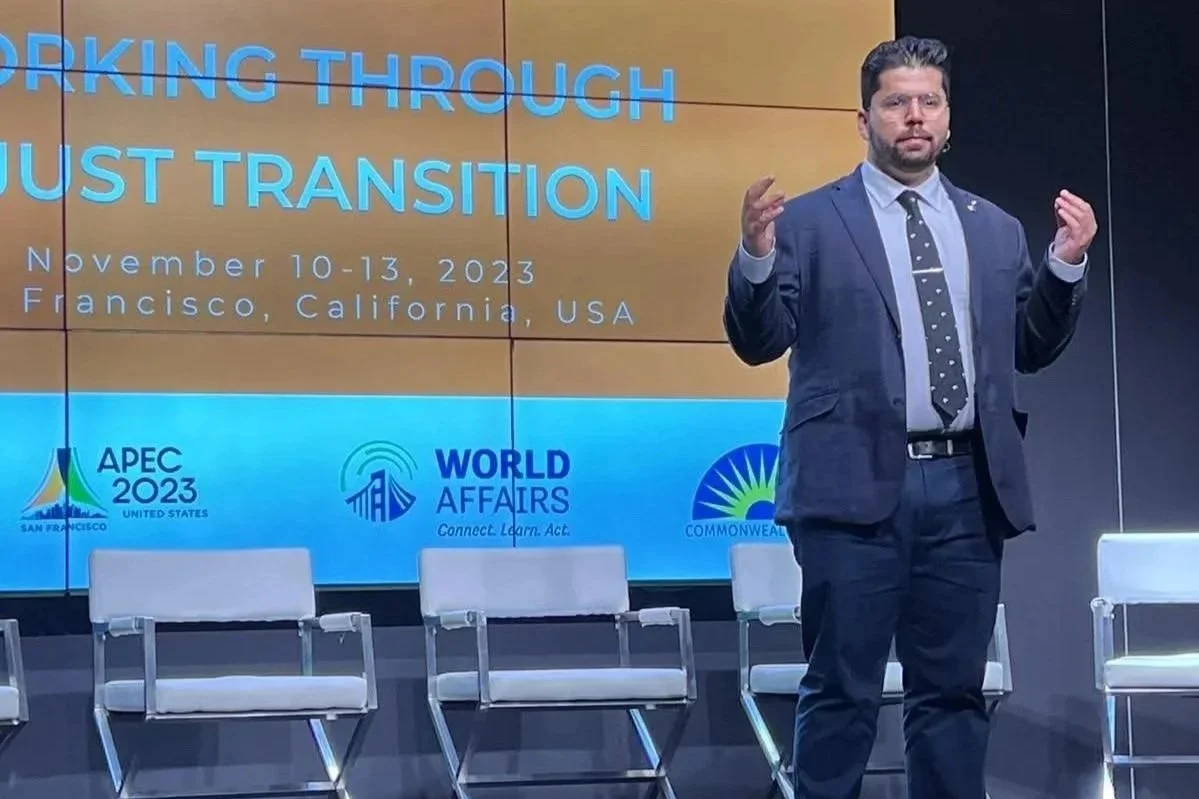 Man in a suit speaking at a conference stage with a large screen behind him displaying the text "WORKING THROUGH TRUST TRANSITION" and event details, with chairs in the foreground.