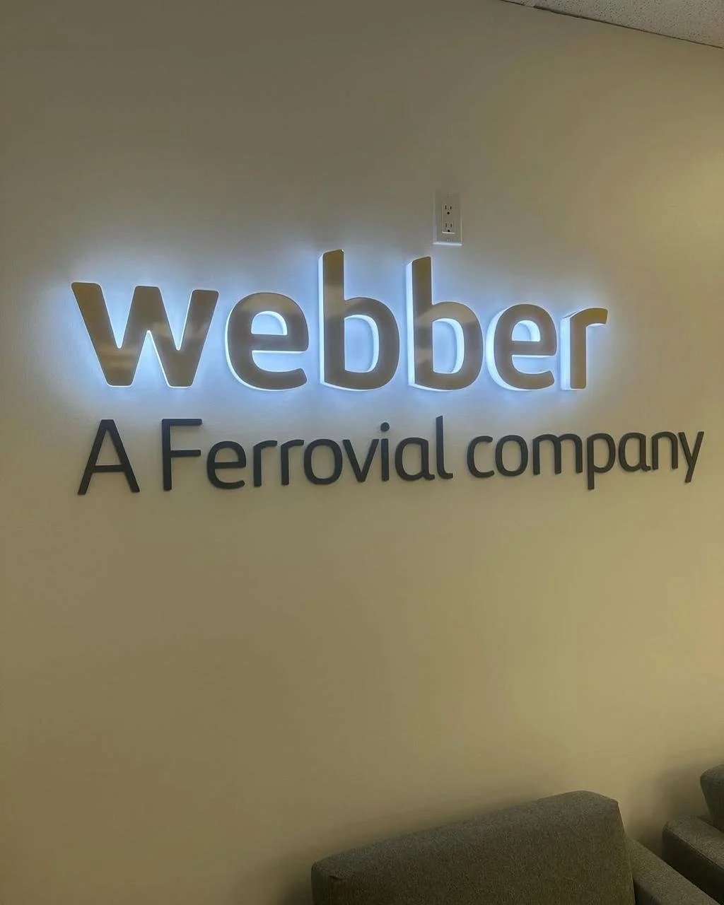 The image shows a wall sign with the company name "Webber" illuminated by backlighting. Below it, there is a tagline that says "A Ferrovial company." There are some chairs at the bottom of the image.
