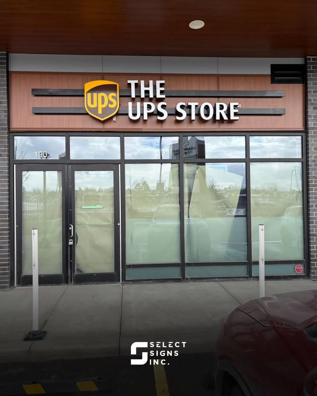 Front view of a UPS store with glass windows and door, and a wooden sign with the UPS logo and the words 'The UPS Store' above the entrance, located at 190. Reflection of the surroundings can be seen on the glass.
