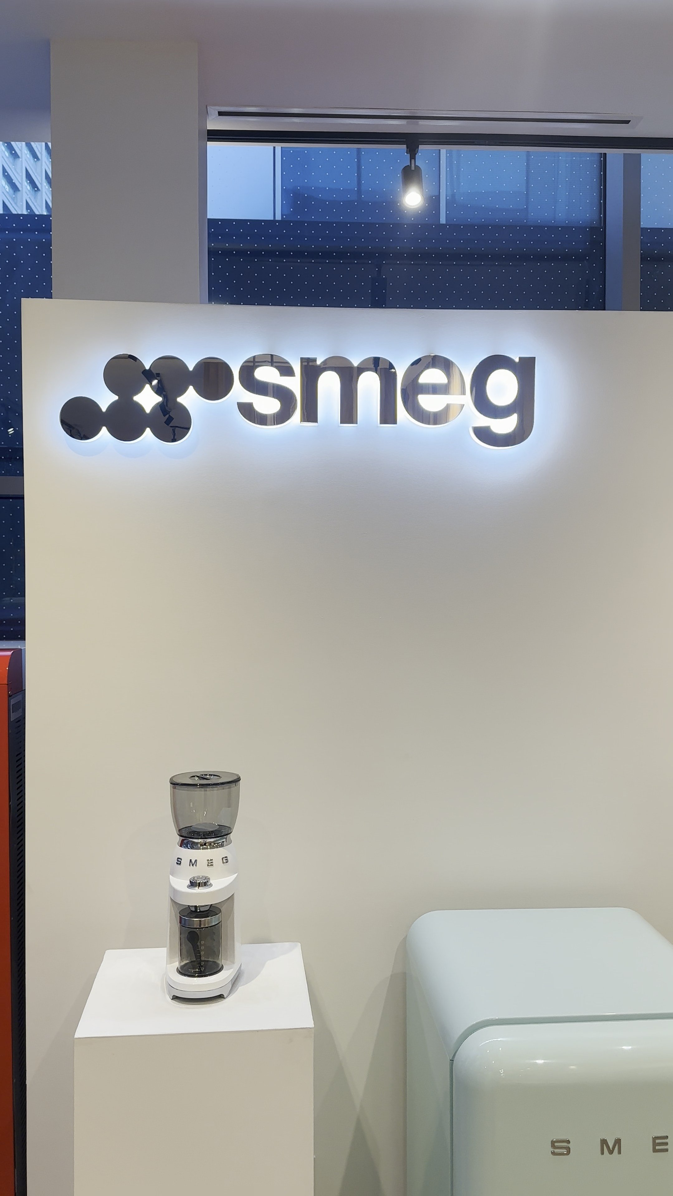 A white Smeg water filter pitcher on a white display stand in front of a white wall with a illuminated 'smeg' logo. The background has a modern interior with large windows showing a cityscape.