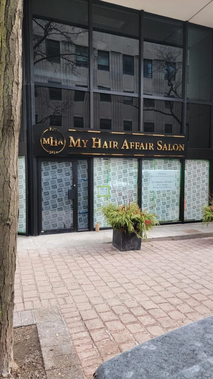  storefront with a sign that reads "My Hair Affair Salon" and two large potted plants outside, with the building's reflective glass windows showing trees and neighboring buildings.