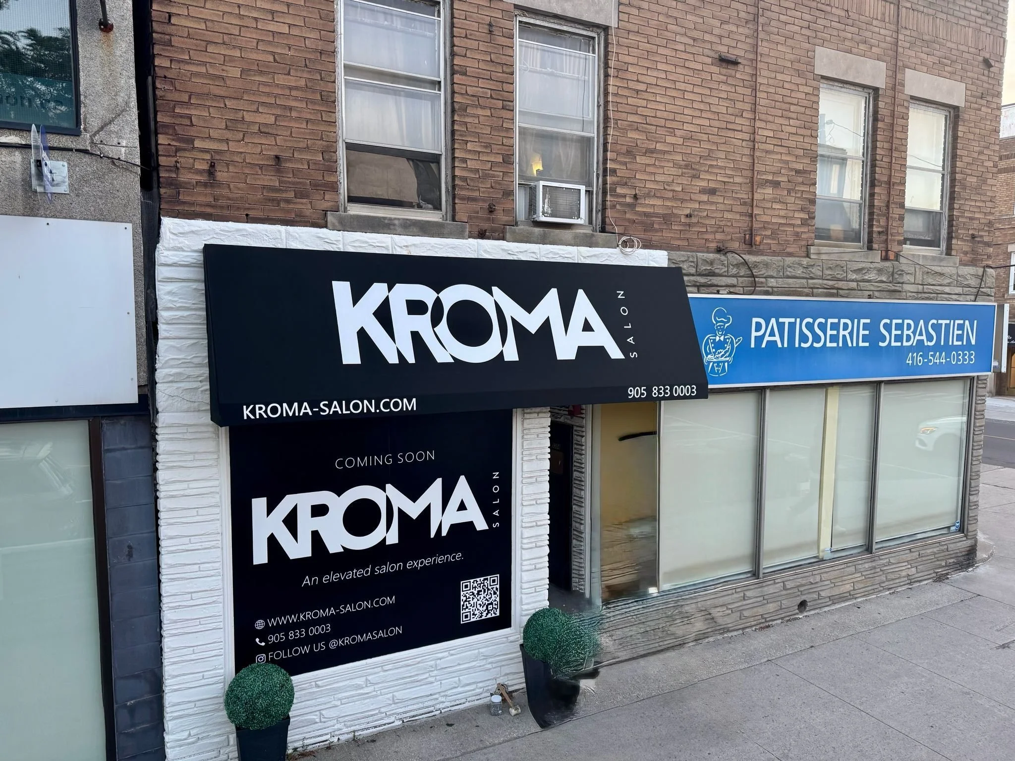 Business storefront with a black sign for Kroma Salon and a blue sign for Patisserie Sebastien, featuring contact information and social media handles. The building has a brick exterior with three windows, and there are small potted decorative shrubs