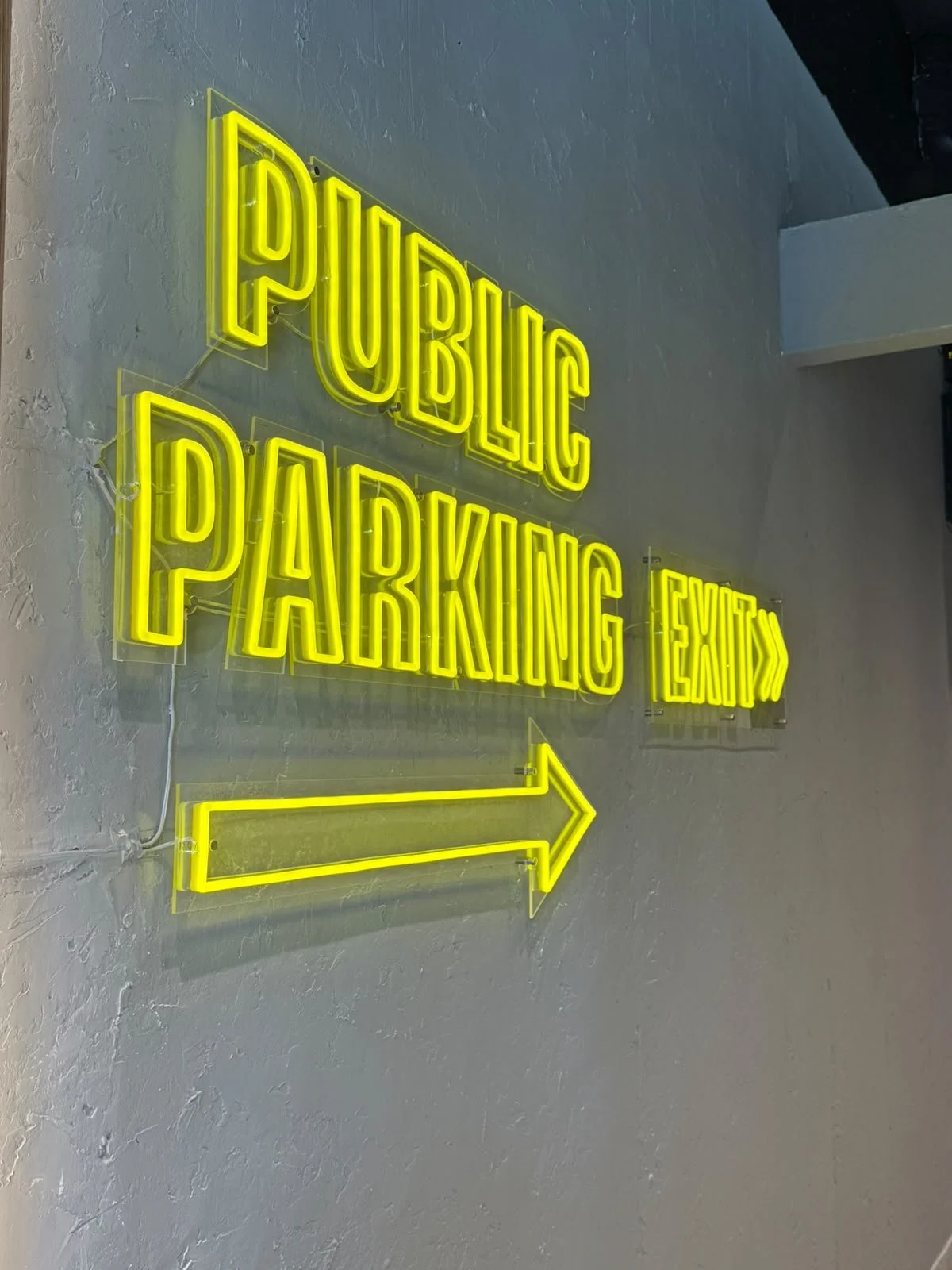 Neon sign on a gray textured wall that reads "PUBLIC PARKING LOT" with an arrow pointing to the right.