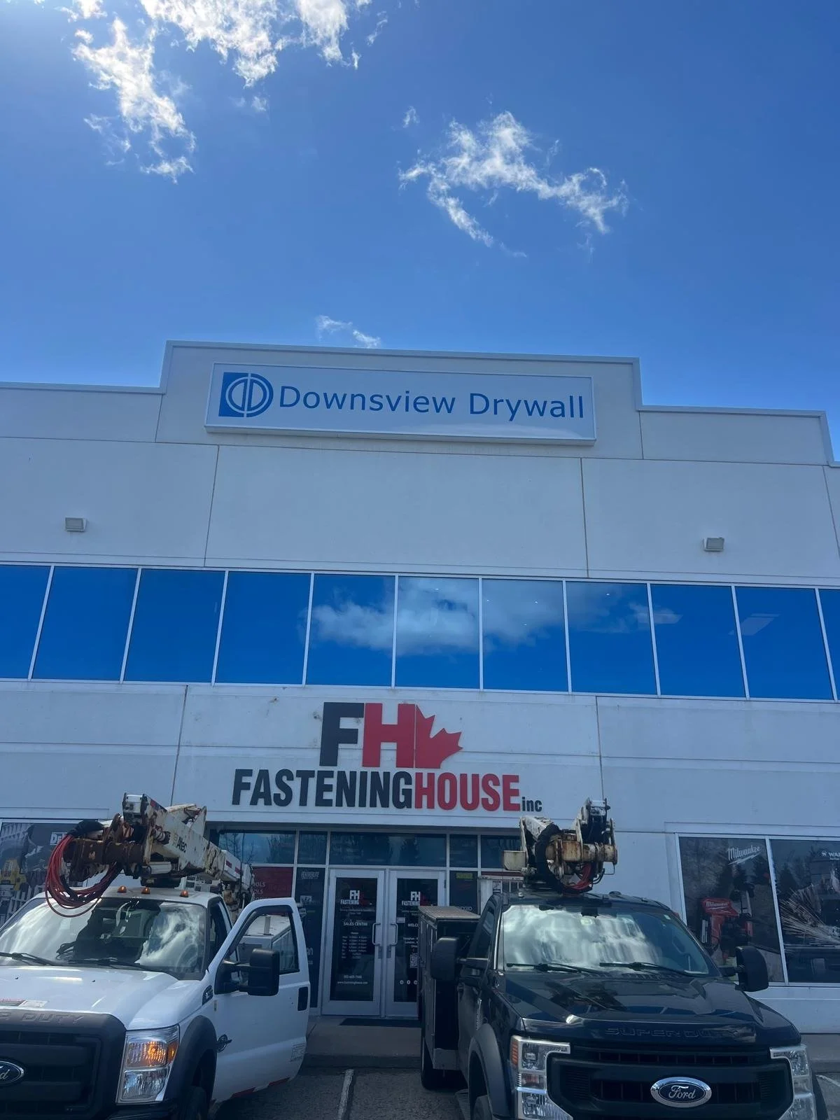 Exterior view of a commercial building with signs for Downsville Drywall and Fastening House Inc., with trucks parked in front on a sunny day with some clouds in the sky.