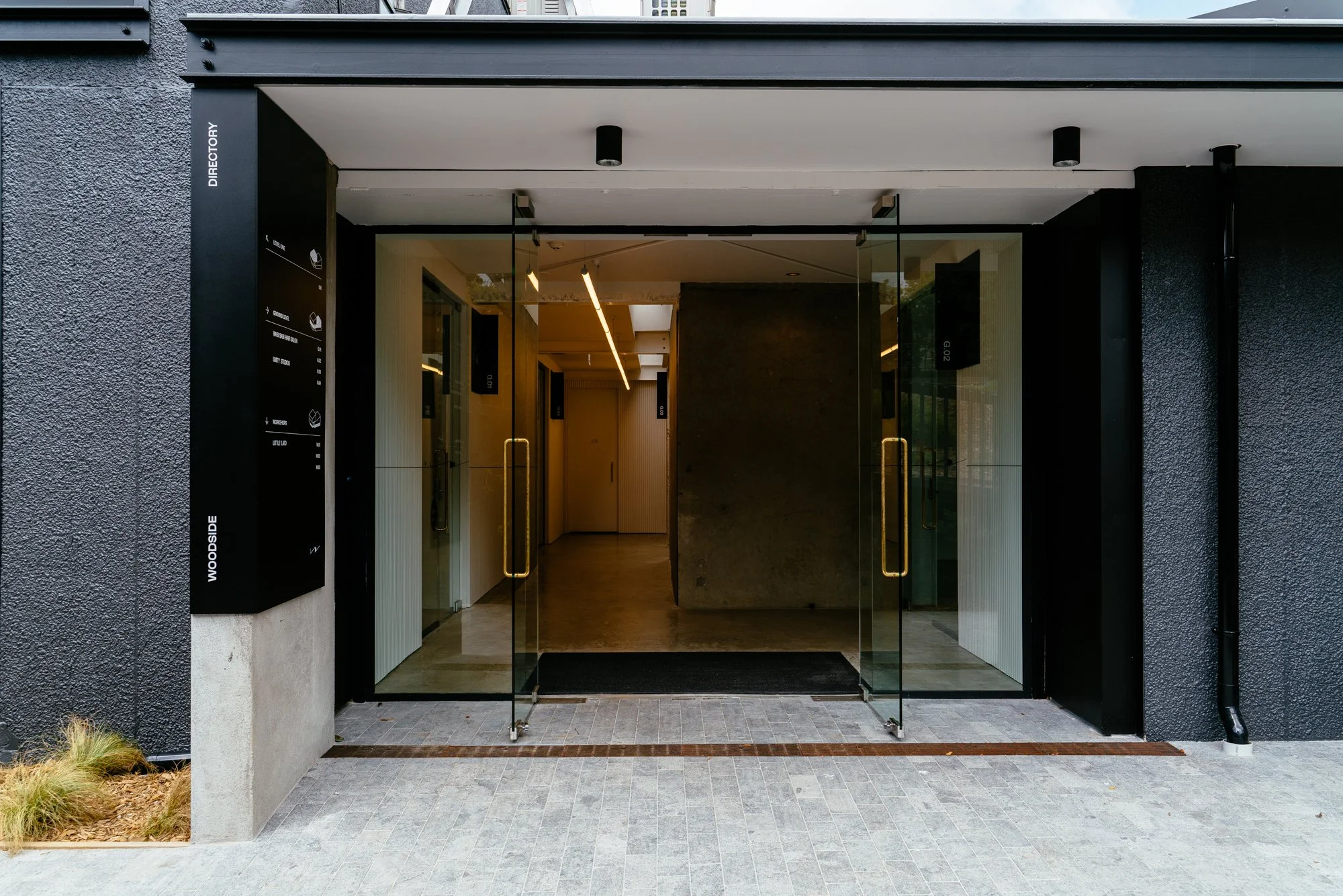 Modern building entrance with glass doors, black exterior framing, and digital directory sign to the left; interior features warm lighting and minimal decor.