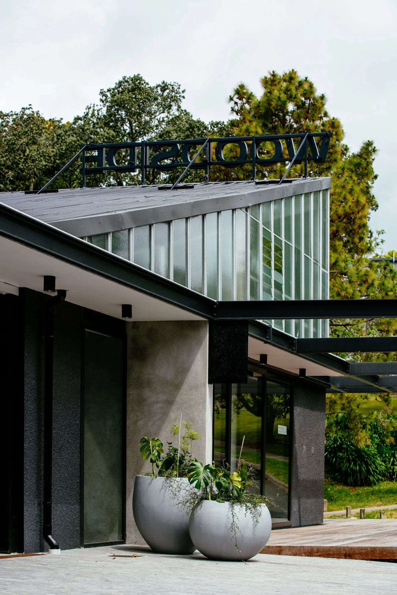 Modern building with glass and metal exterior, large white planters with green plants, and trees in the background.