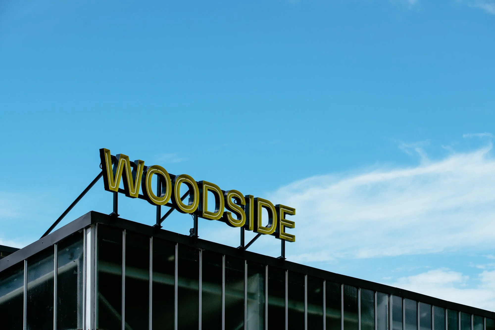 The image shows the top part of a modern building with large glass windows and an illuminated yellow sign that says "WOODSIDE" on the rooftop, against a blue sky with some clouds.