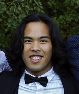 Close-up of a young man with long black hair, wearing a tuxedo with a bow tie, smiling outdoors with greenery in the background.