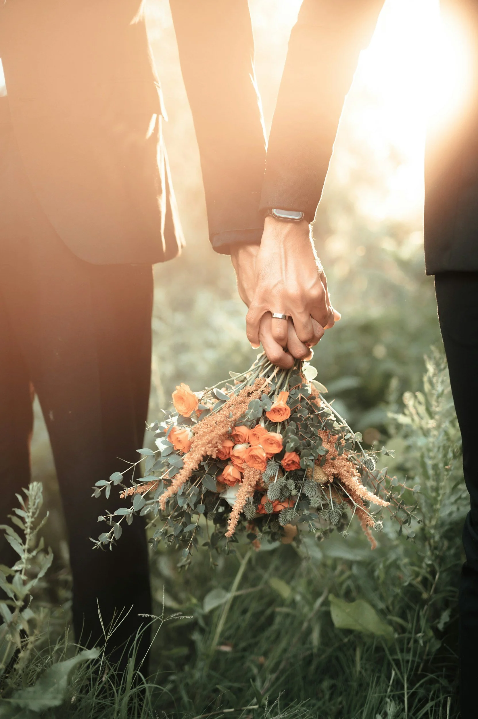 A couple holding hands outdoors in a field, with the woman holding a bouquet of flowers.
