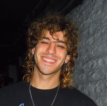 A young man with curly hair, earrings, and a necklace smiling in front of a brick wall.