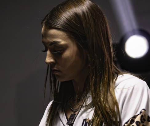 A woman with long brown hair and a serious expression, wearing a white shirt, indoors with a blurred background.
