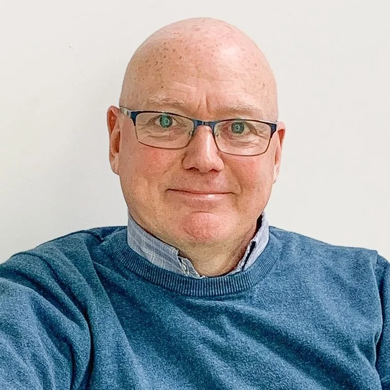 A friendly and approachable male counsellor with glasses against a plain white wall. He is wearing a blue sweater over a collared shirt.