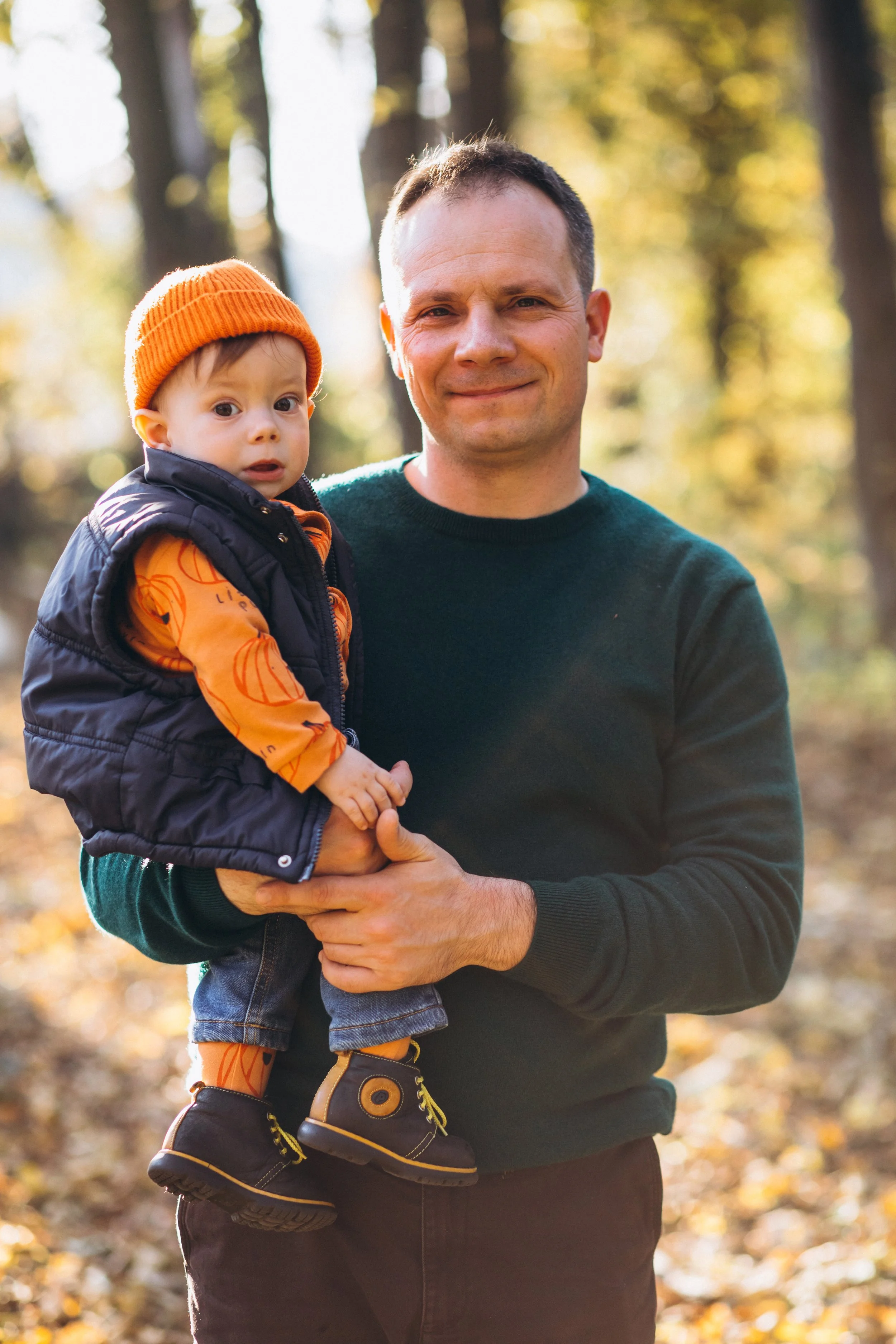A man holding a young child outdoors in a wooded area during autumn, with trees and fallen leaves in the background.