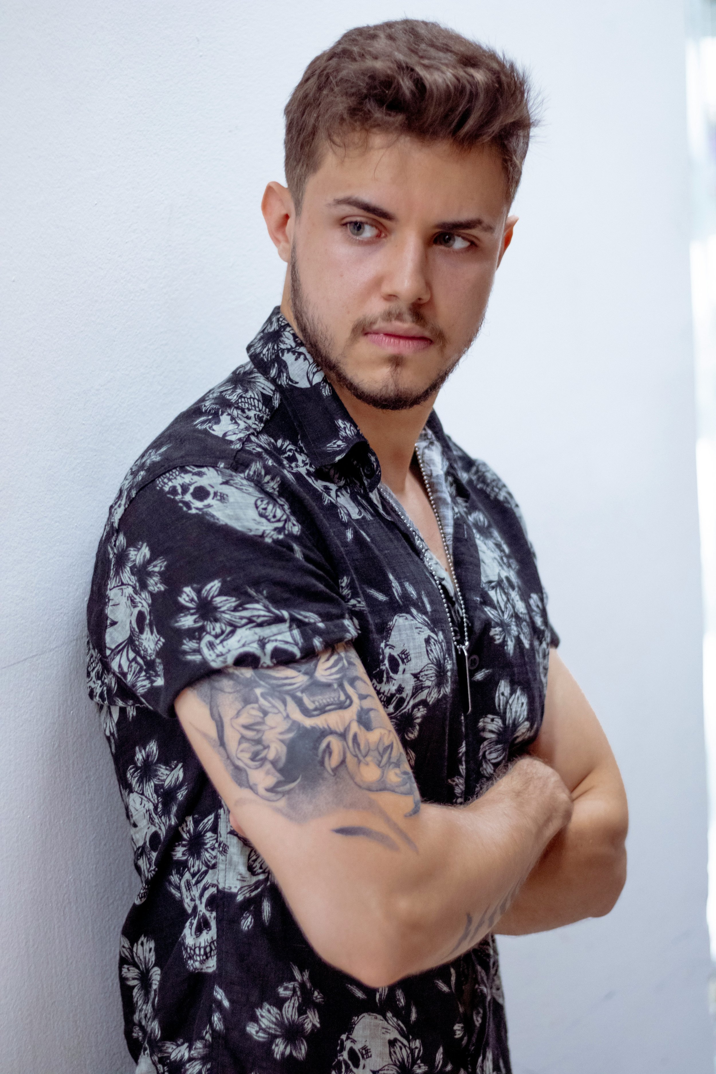 Young man with short brown hair and a beard, wearing a black short-sleeved shirt with white floral and skull patterns, standing with arms crossed against a white wall, looking to the side.