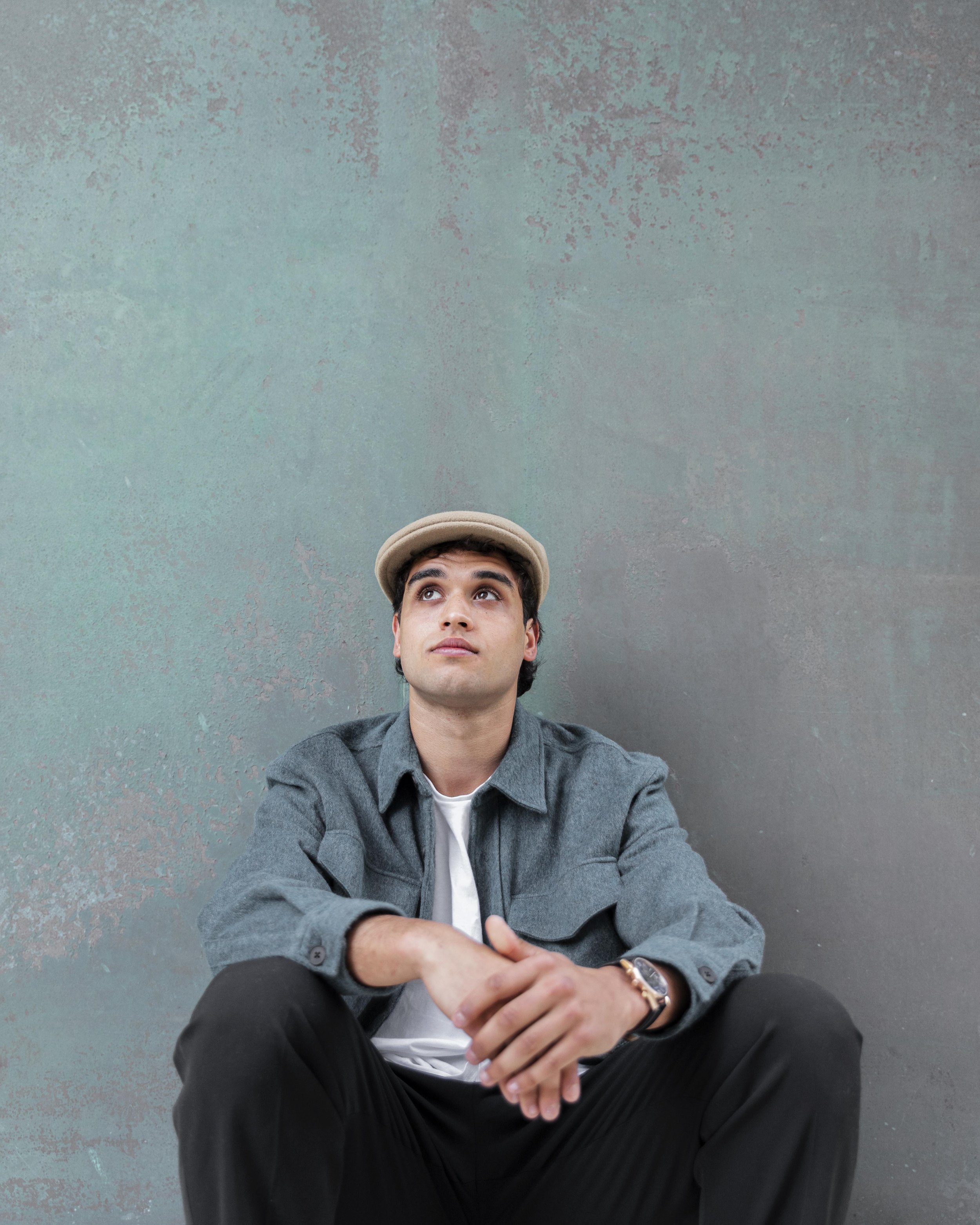 A young man with dark hair and light skin tone sitting on the ground, wearing a gray shirt, black pants, a beige cap, and a wristwatch, looking up with a neutral expression against a textured teal wall.
