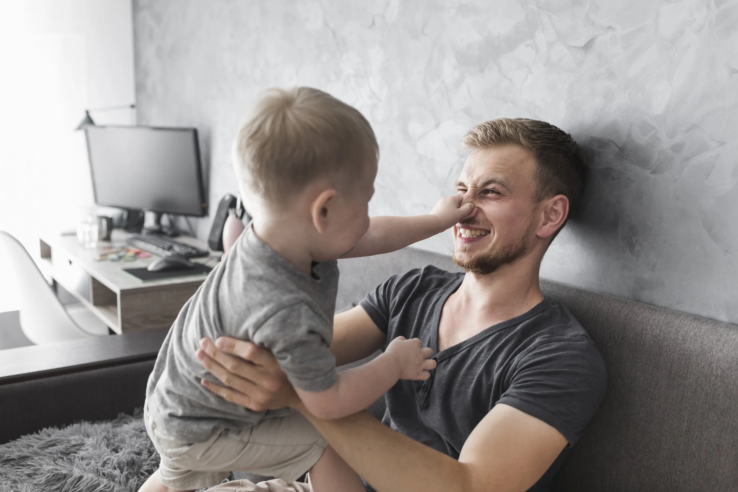 A young man and a toddler playing together indoors. The toddler is reaching to touch the young man's nose, and they are smiling and laughing.
