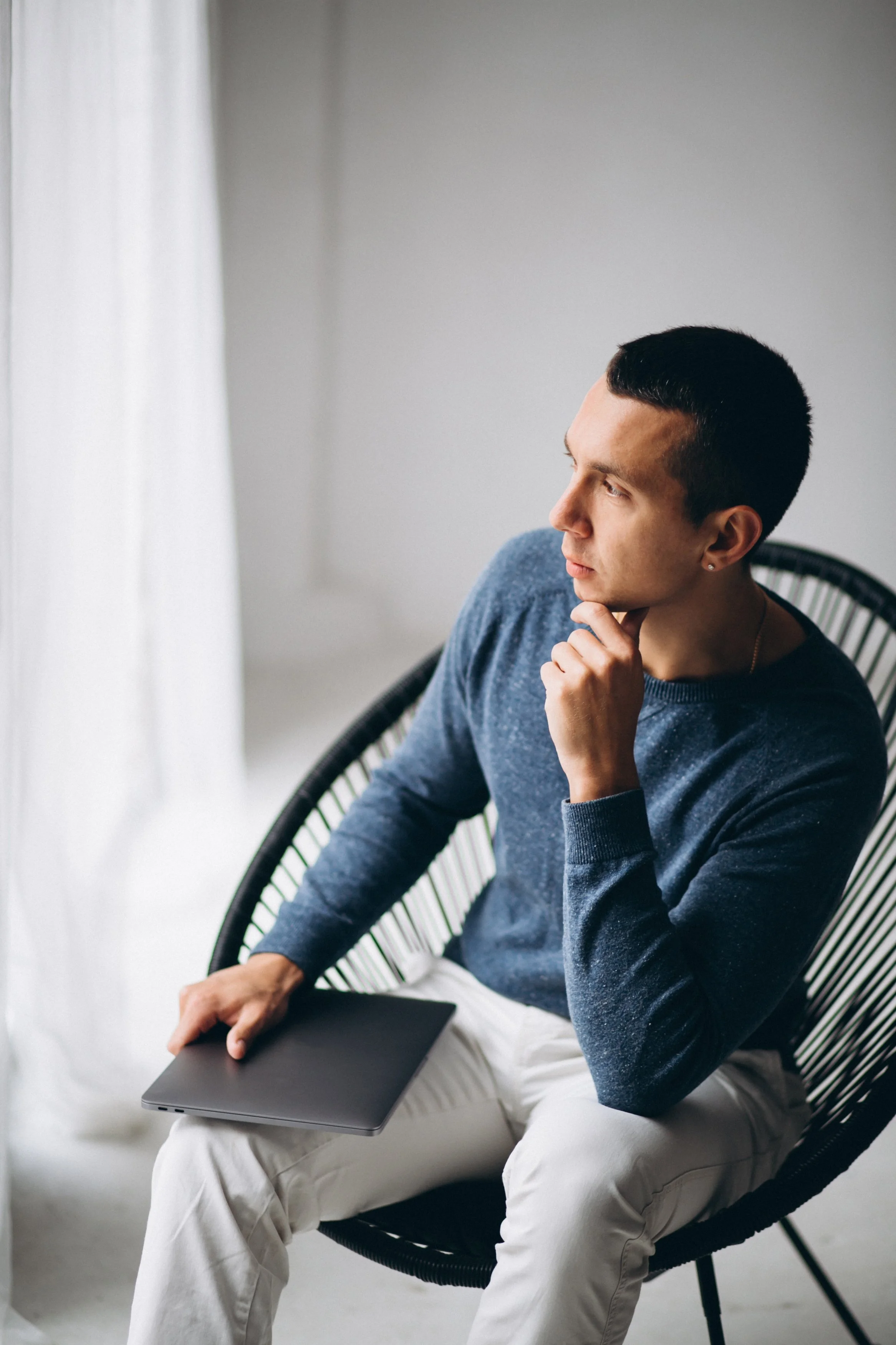 A young man with short dark hair, wearing a blue sweater and white pants, is sitting in a black rattan chair near a window with white curtains, holding a closed laptop on his lap and resting his chin on his hand, looking thoughtfully out the window.