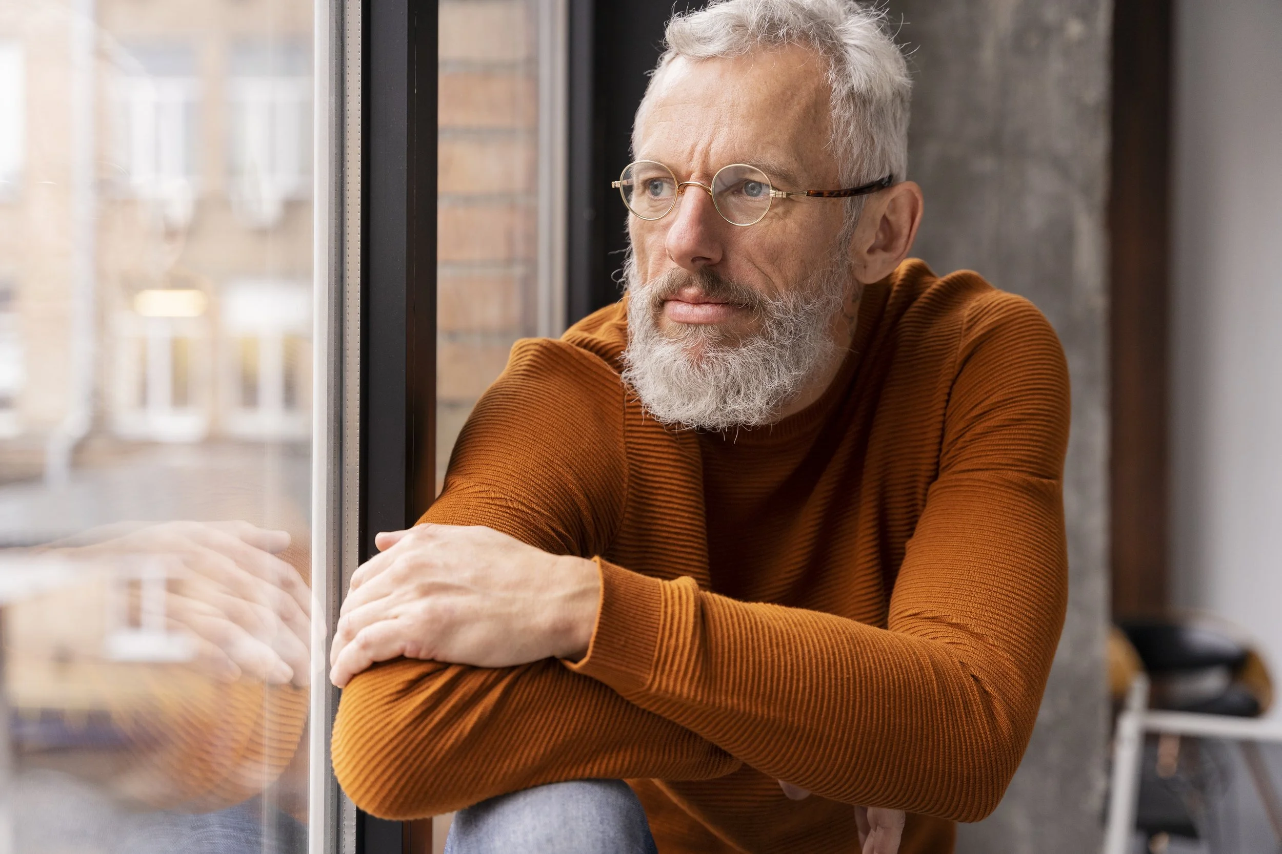 Older man with gray hair, glasses, a beard, and a mustache, wearing an orange sweater, sitting by a window, looking out contemplatively.