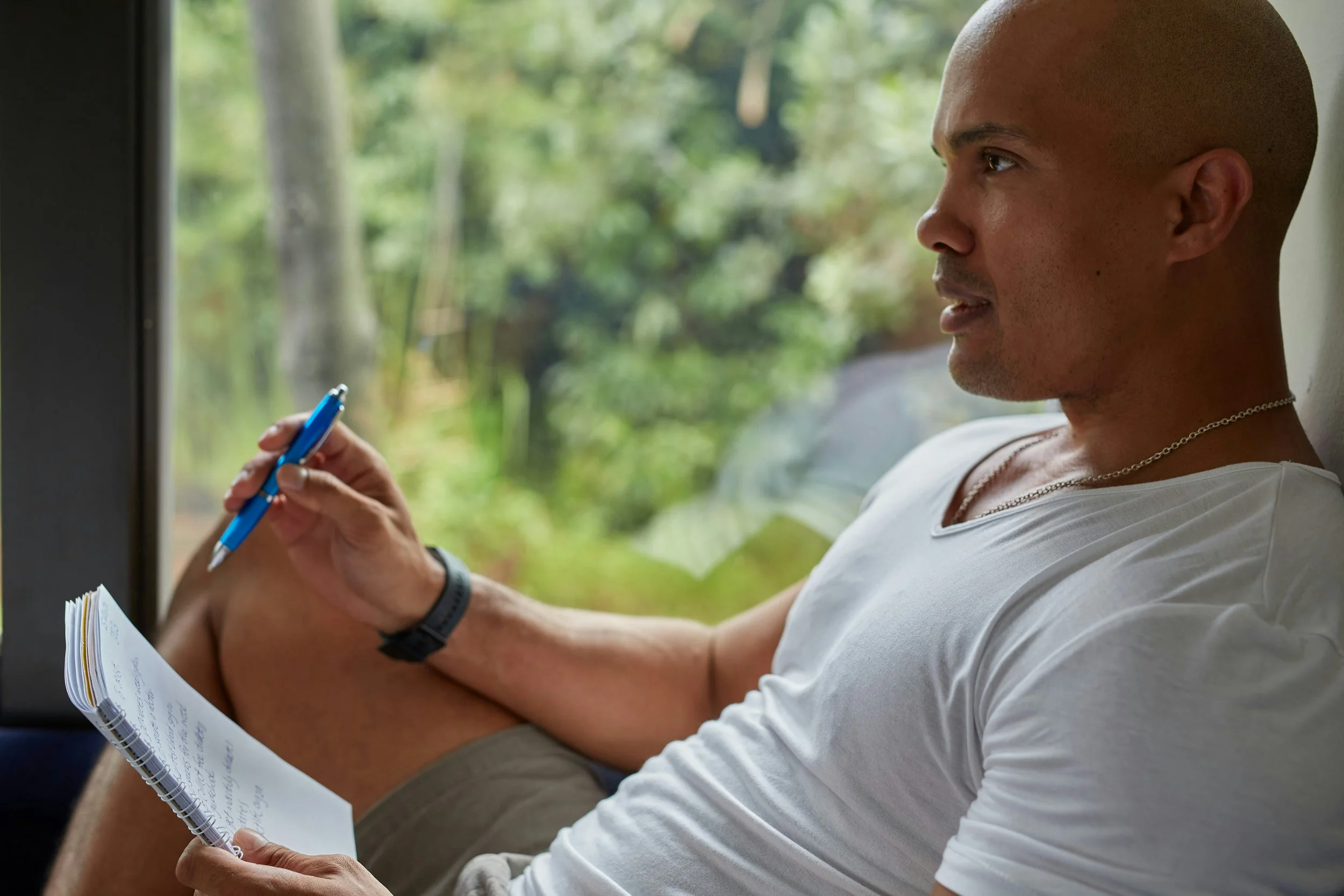 Man sitting by a window, reading notes and writing with a pen, with greenery outside