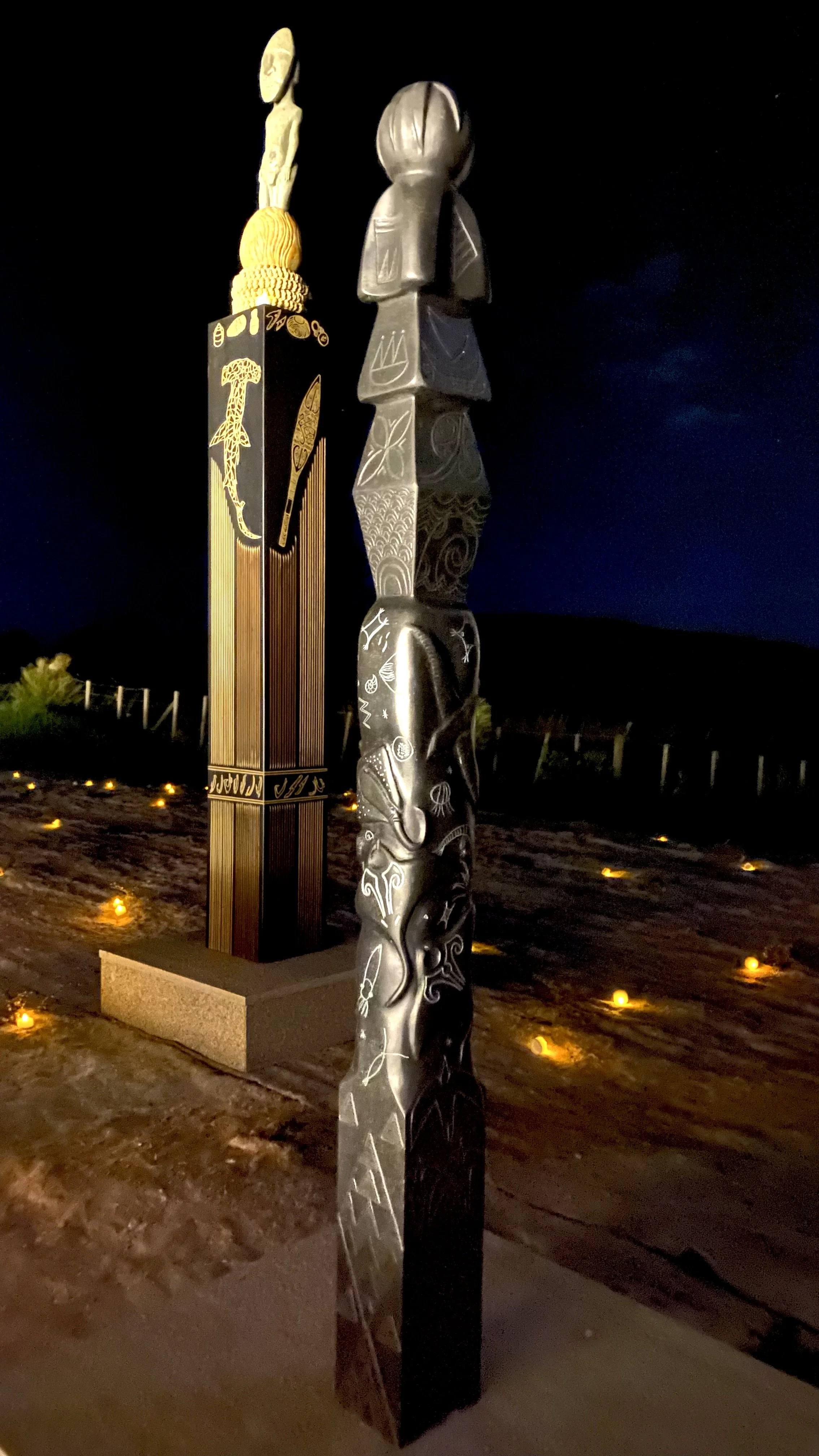 Nighttime view of carved wooden and stone ancestral totem poles with intricate designs, lit by small ground lights, set against a dark sky.