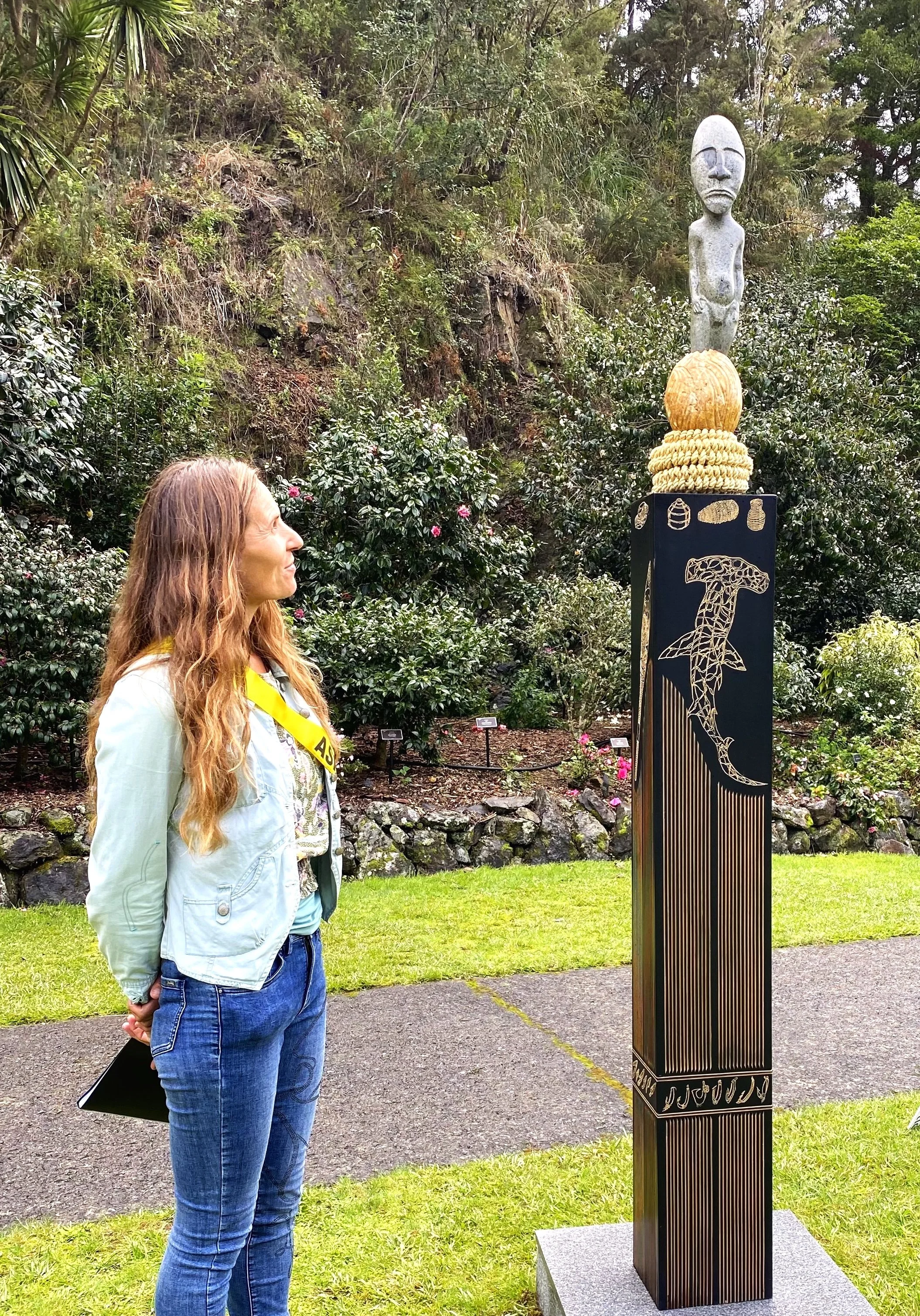 A woman with long hair, denim jacket, and jeans stands outdoors admiring a sculpture on a tall black pedestal, surrounded by green bushes and trees.