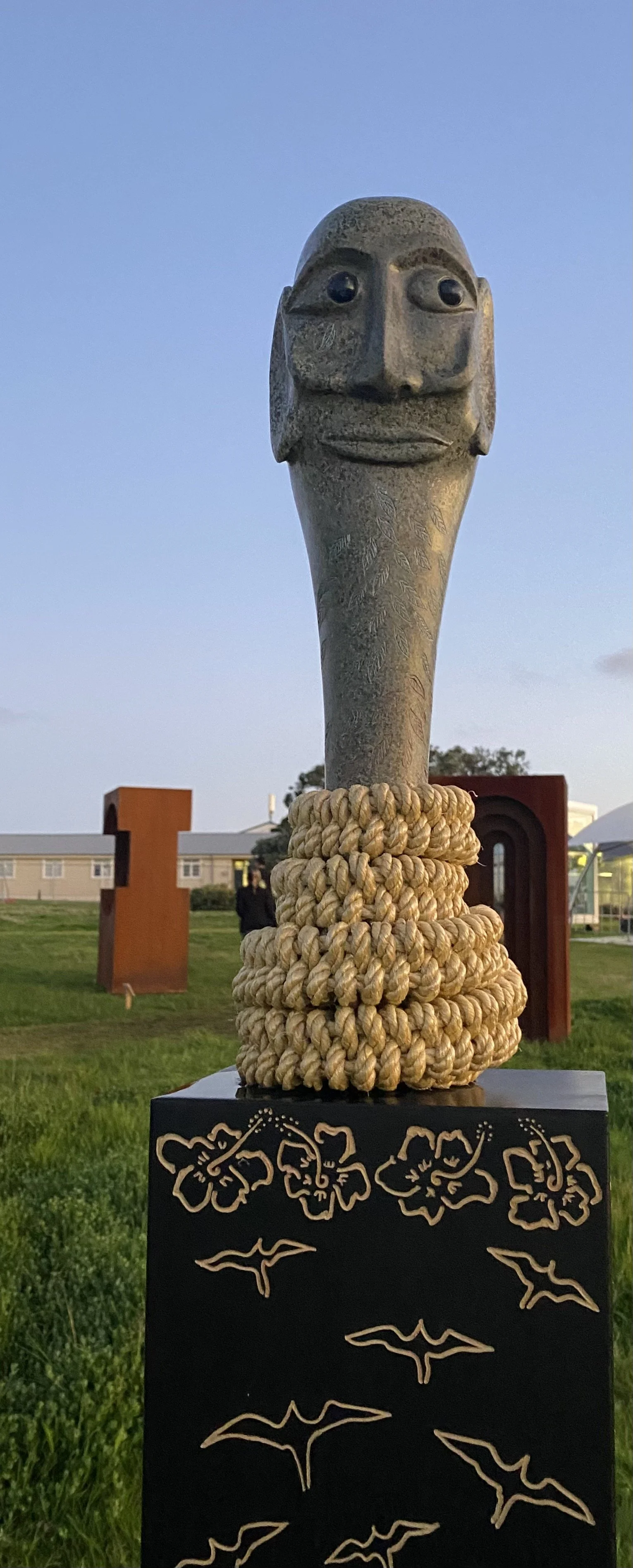 A stone sculpture with a stylized human face, featuring large eyes and a subtle smile. It is mounted on a black pedestal decorated with floral and bird motifs. The sculpture has a series of ropes wrapped around its lower section. The background shows grass and a structure.