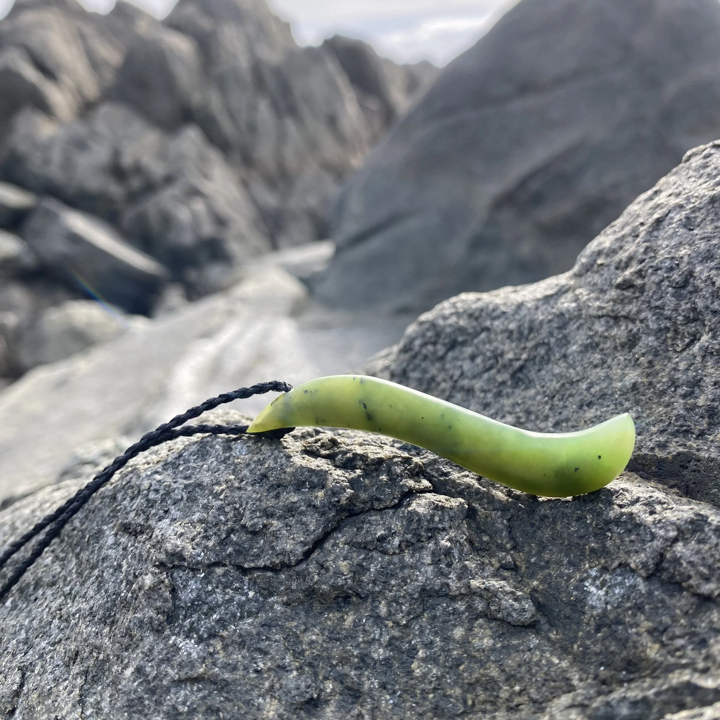 Close-up of a contemporary pounamu greenstone Maori necklace shape with black string on gray rocks in a rocky oceanside landscape.