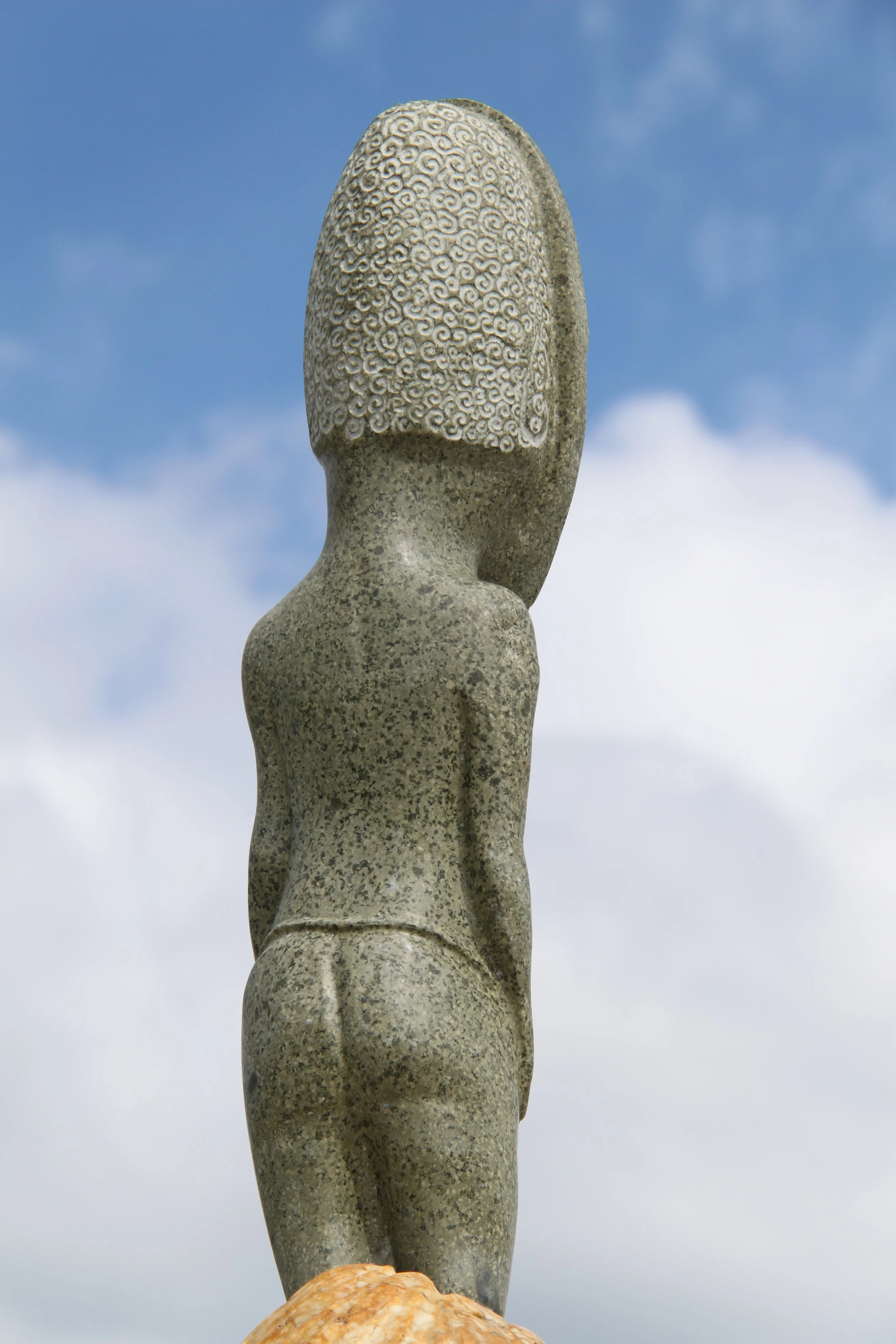 Stone statue of a human figure with a patterned head against a blue sky.
