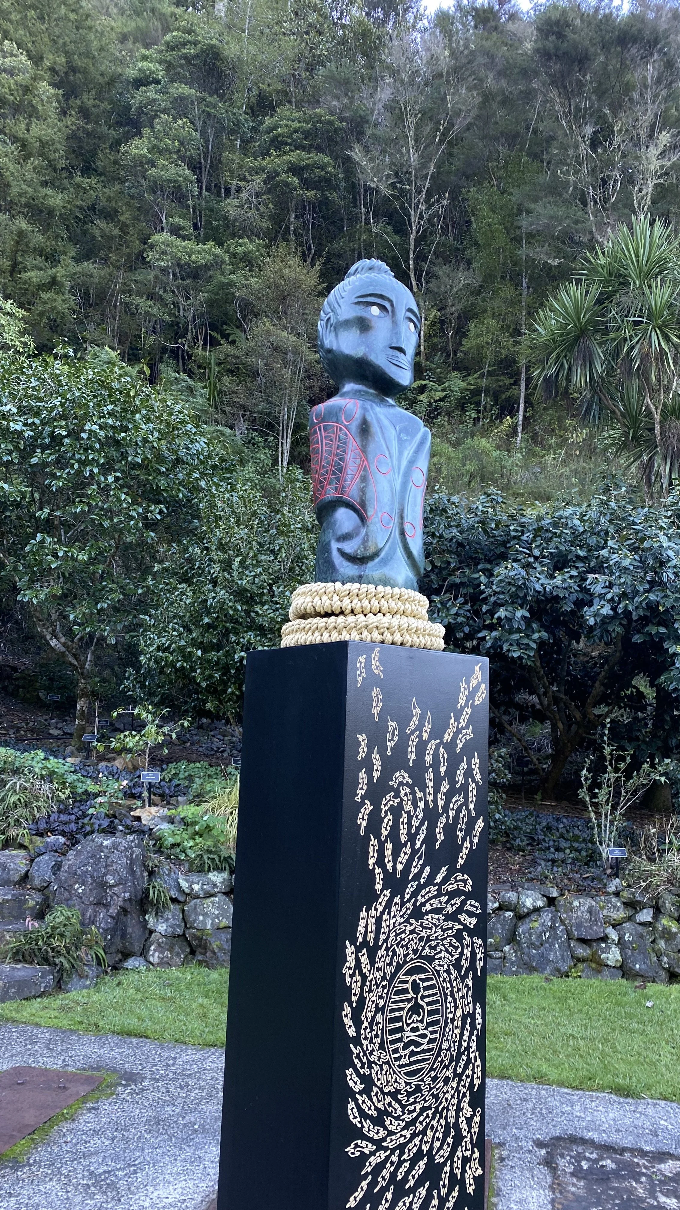A sculpture of a person, an ancestor of Polynesian origins carved in Serpentine, with a large Manta ray tattoo as a cloak that features symbols from aroung the Pacific islands, standing upon a pou, or pedestal with intricate carving details.