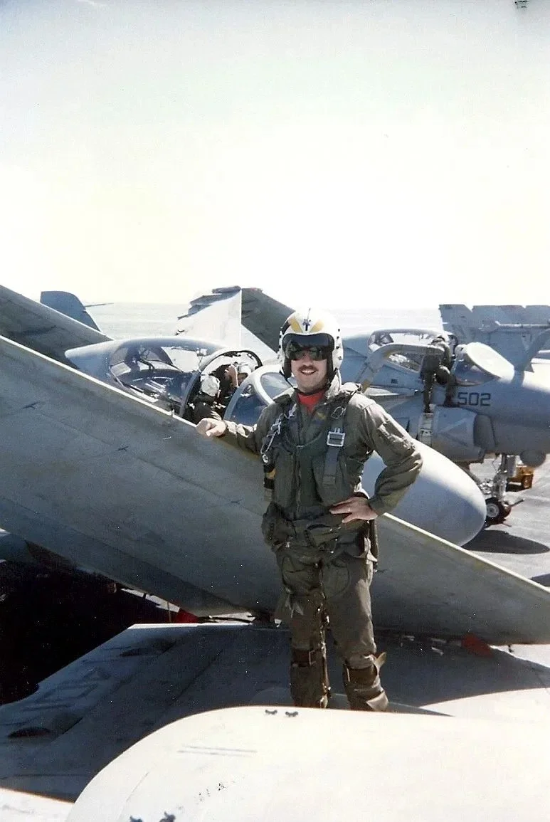 A naval pilot stands on the wing of an aircraft parked on the deck of an aircraft carrier.
