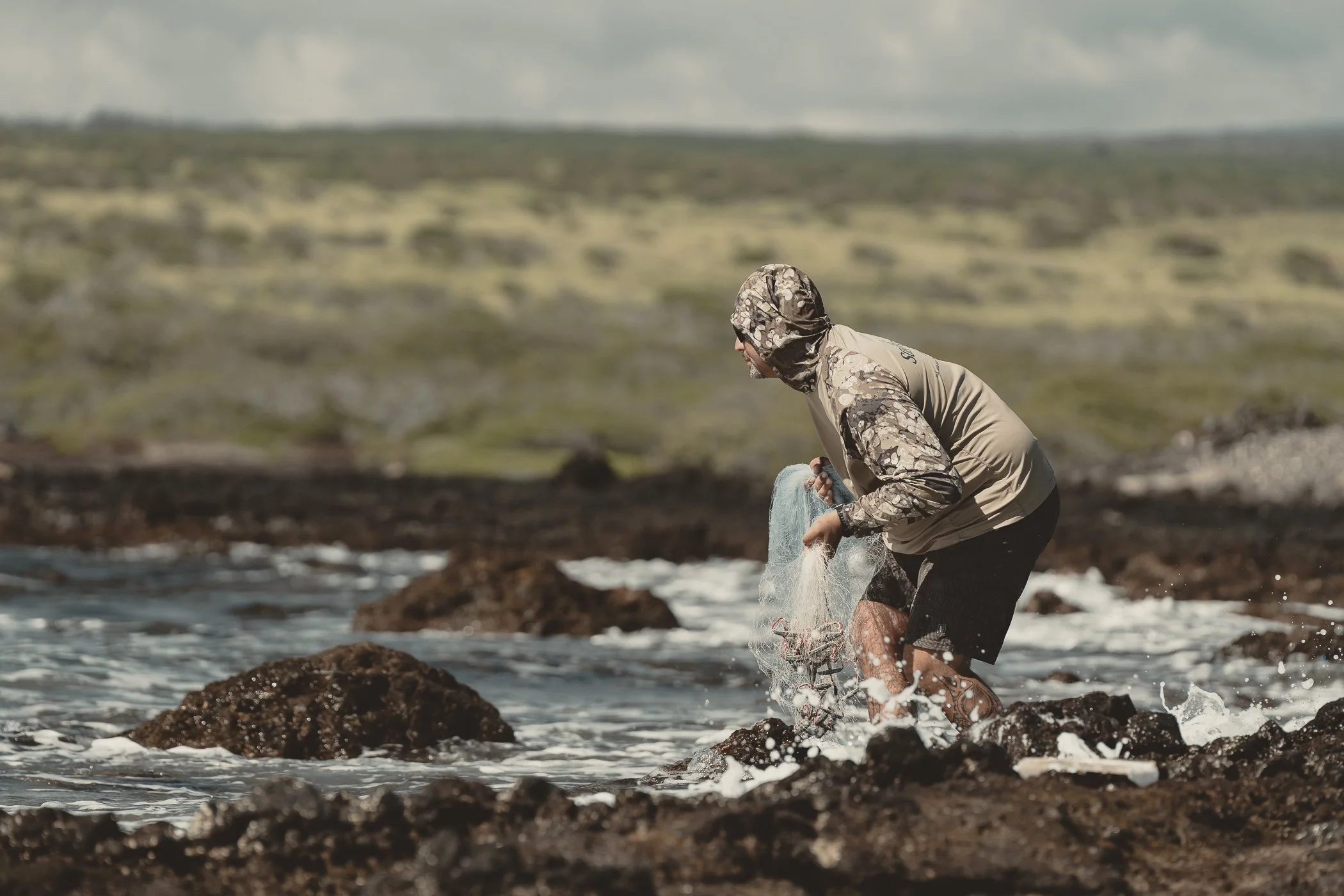 Person wearing a camouflage hooded jacket and black shorts collecting marine debris in the ocean near rocks.
