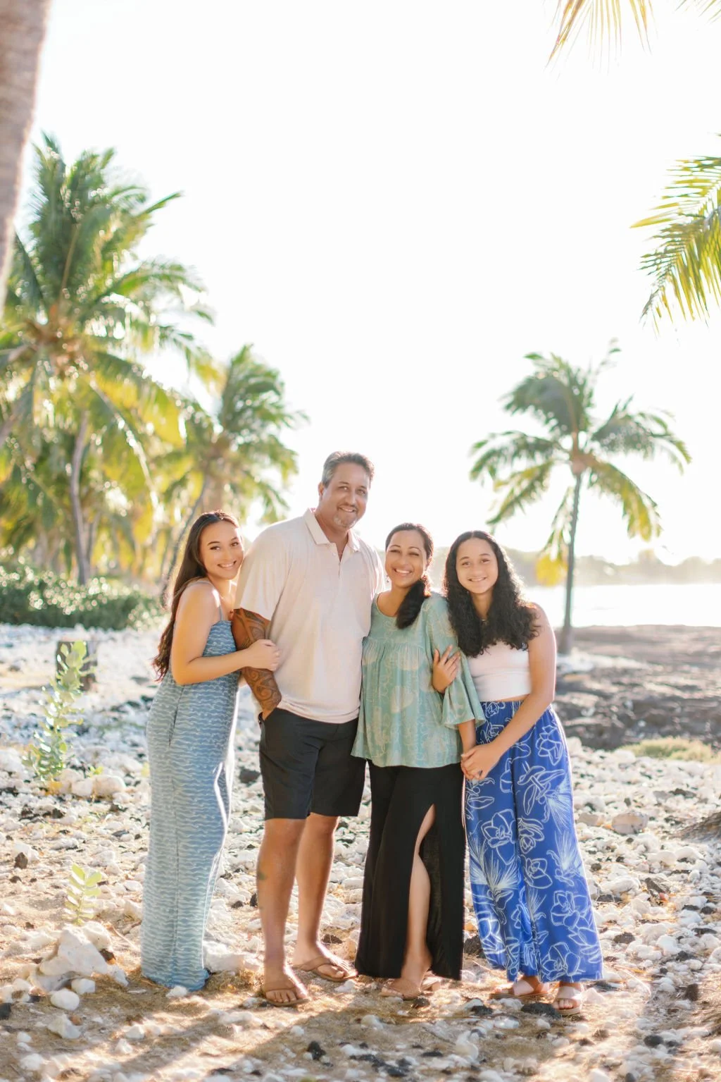 A smiling family of four standing on a rocky beach with palm trees in the background during daytime.