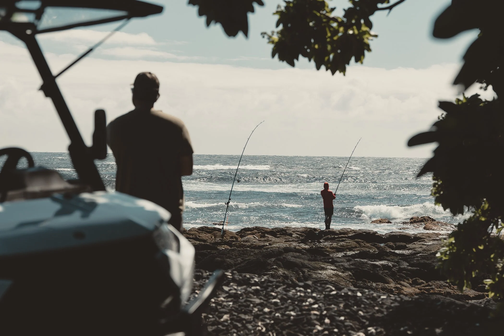 Two people fishing on a rocky beach with a boat and trees in the foreground, ocean waves in the background.