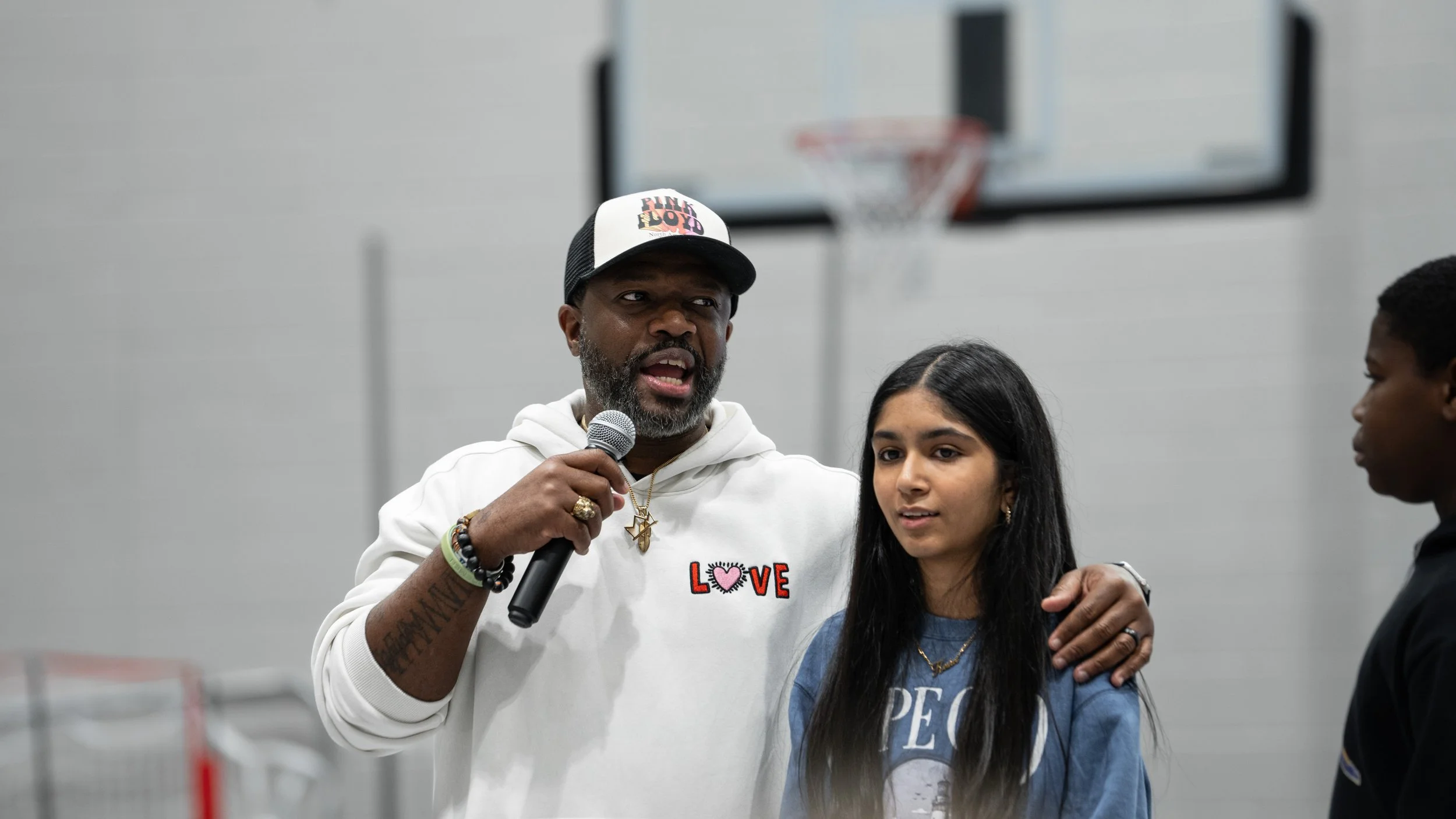 Bobby Gueh with Brothers Making Moves wearing a white hoodie with a 'LOVE' graphic, holding a microphone, and standing next to a young woman with long dark hair, while another person looks on in a gymnasium with a basketball hoop in the background.